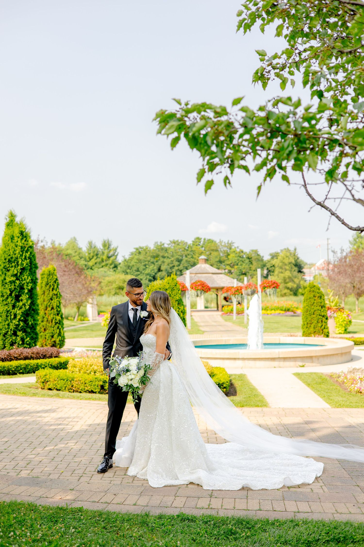 Couple embraces on brick pathway surrounded by sculpted gardens and water features.