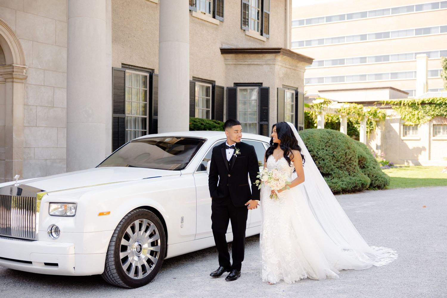 Elegant wedding photo with couple standing by their white Rolls-Royce outside a grand building.