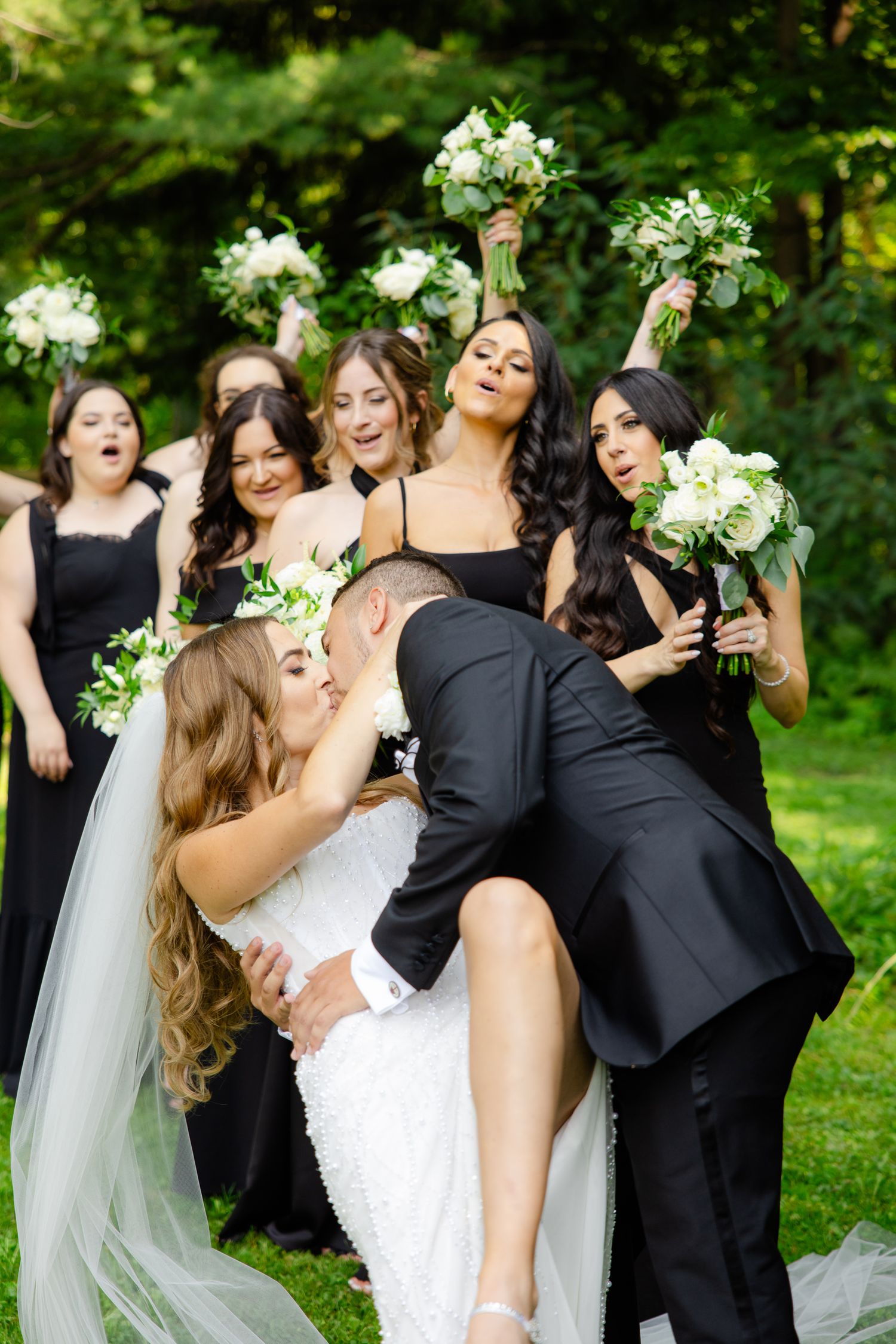 Fun wedding party photo with bridesmaids in black dresses holding white bouquets as couple shares dramatic dip kiss.
