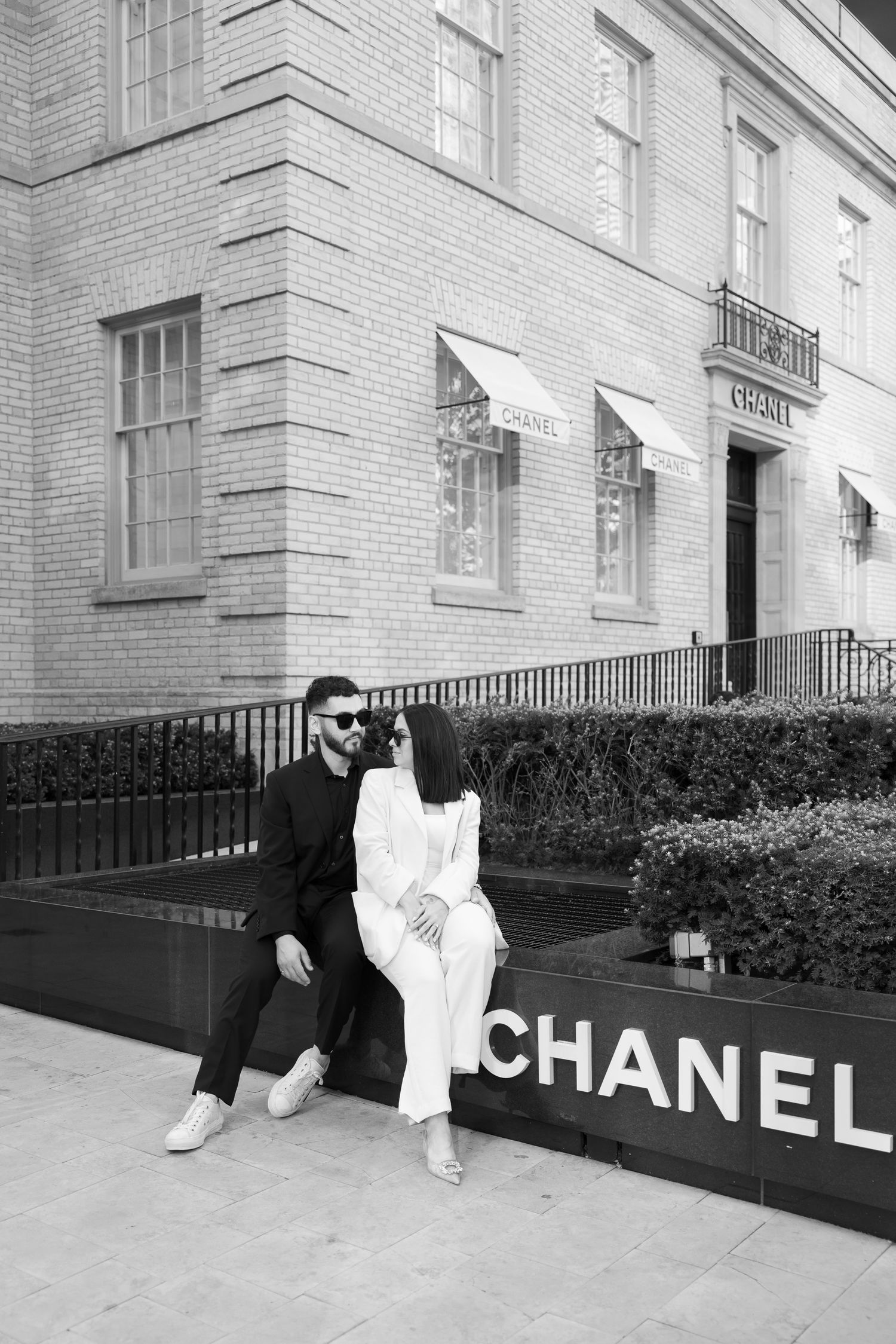 Couple in contrasting black and white outfits posing outside Chanel store entrance.