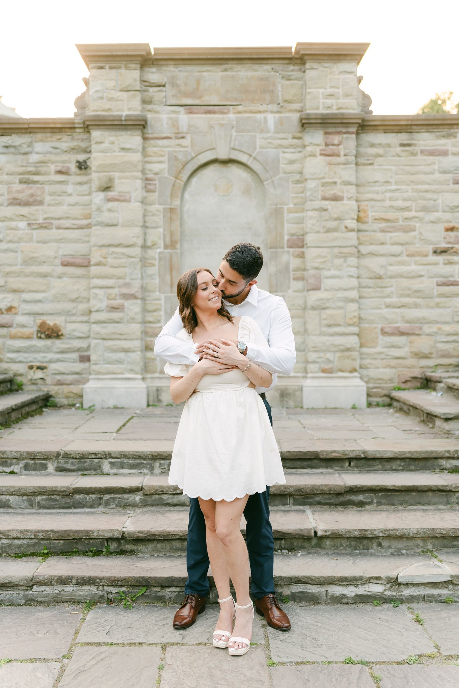 Couple in coordinating white outfits poses on stone steps in front of an architectural archway.