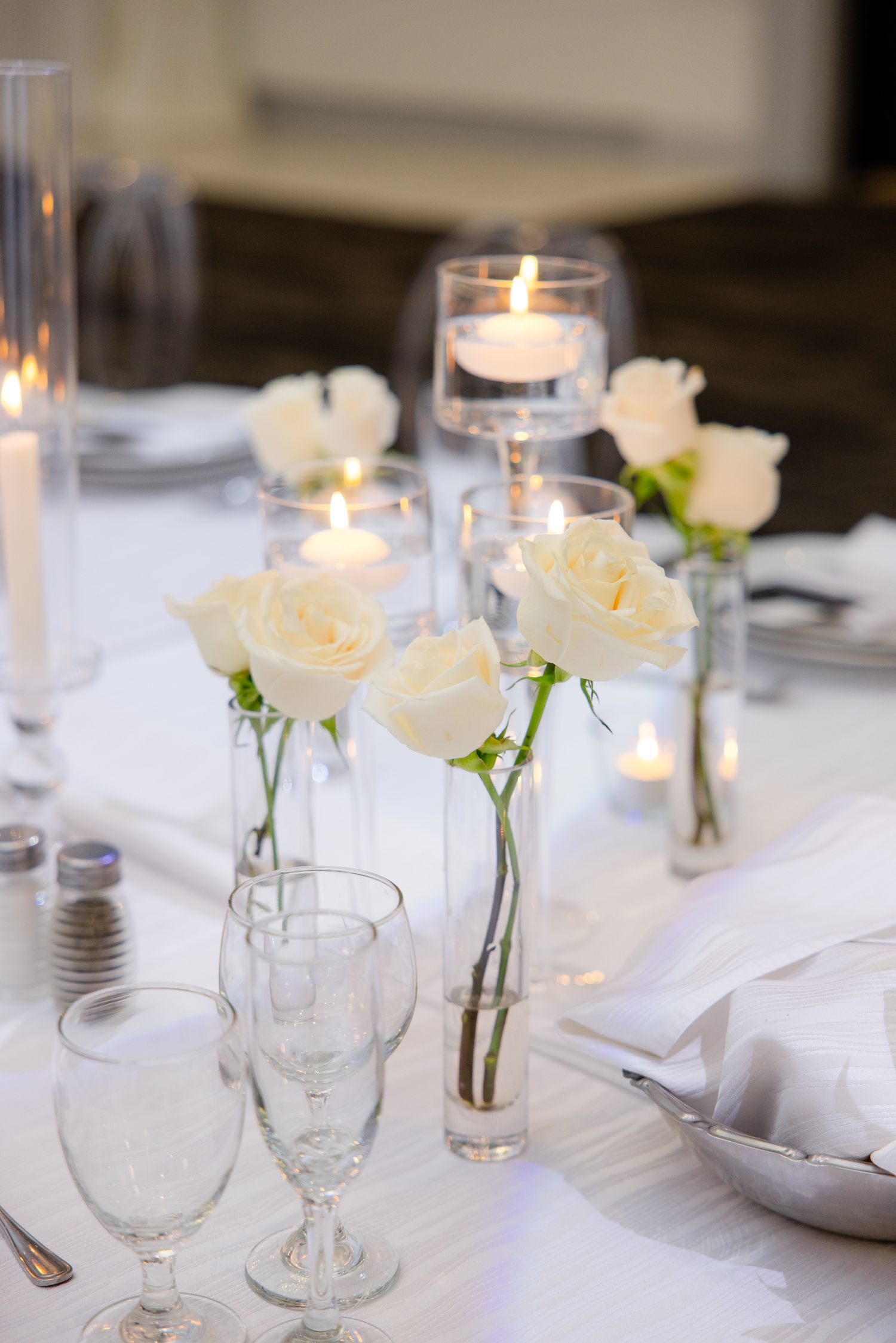 Elegant wedding table setting with white roses in bud vases and glowing candlelight creating romantic ambiance.