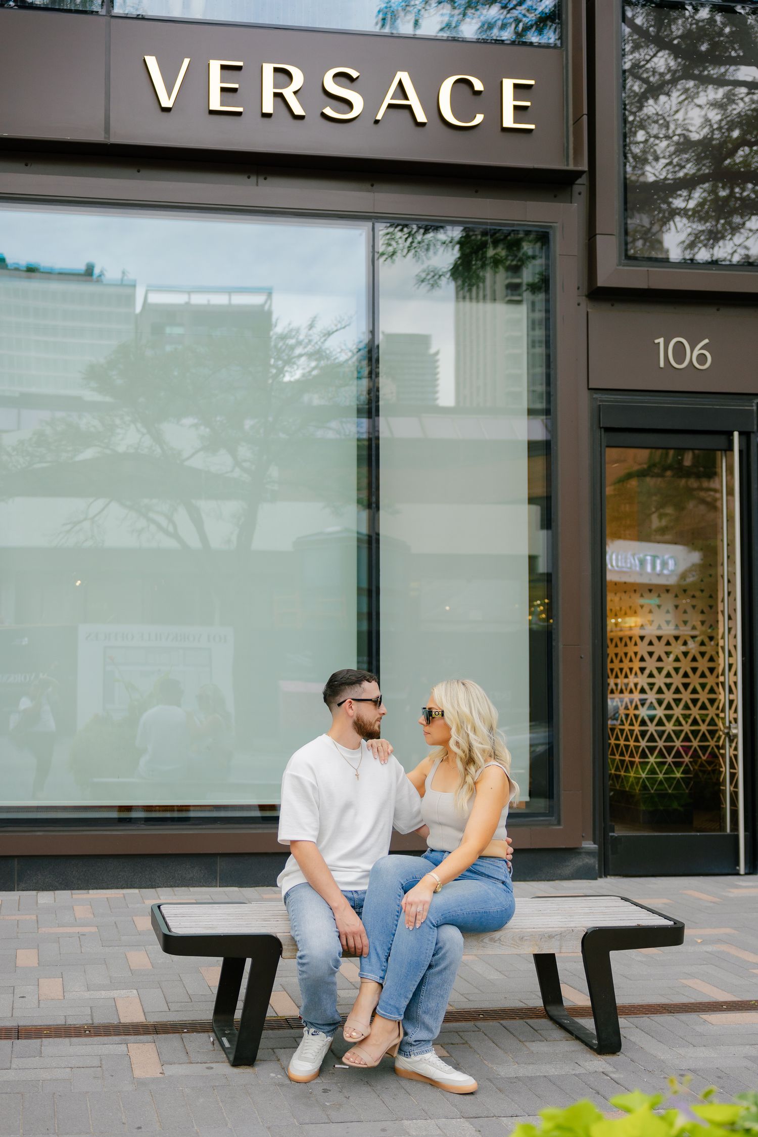Casual couple sitting together on modern bench outside Versace boutique.