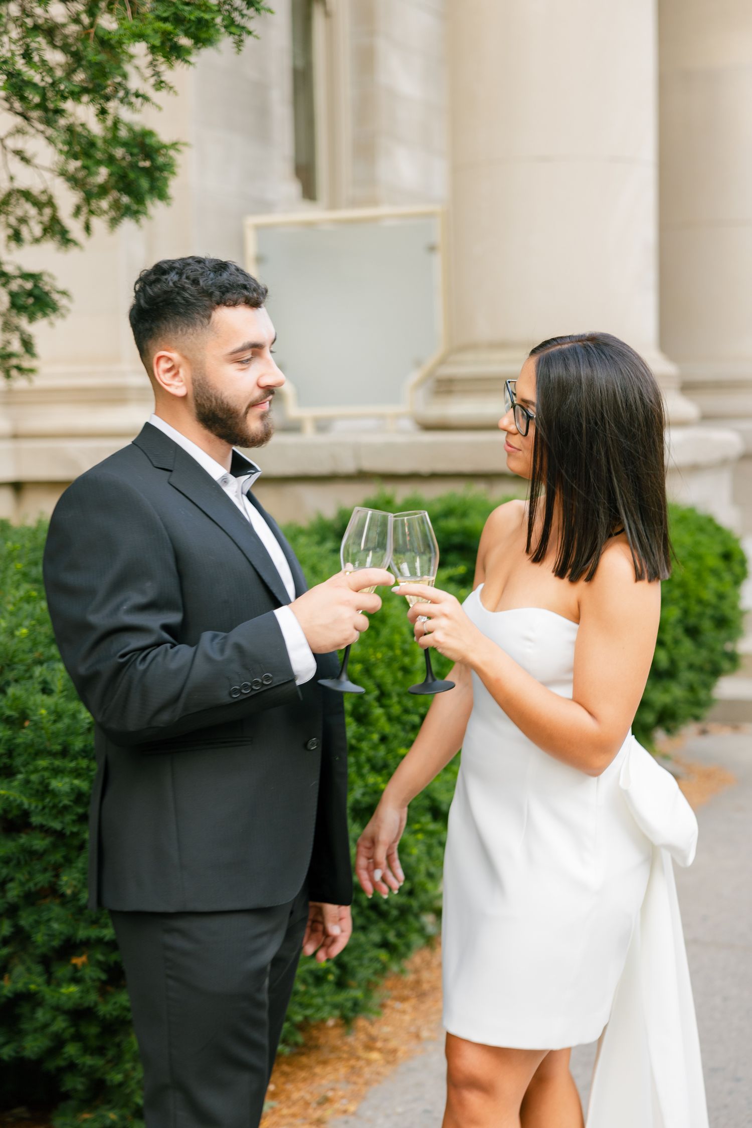 Elegantly dressed couple toasting with wine glasses by a neoclassical building.