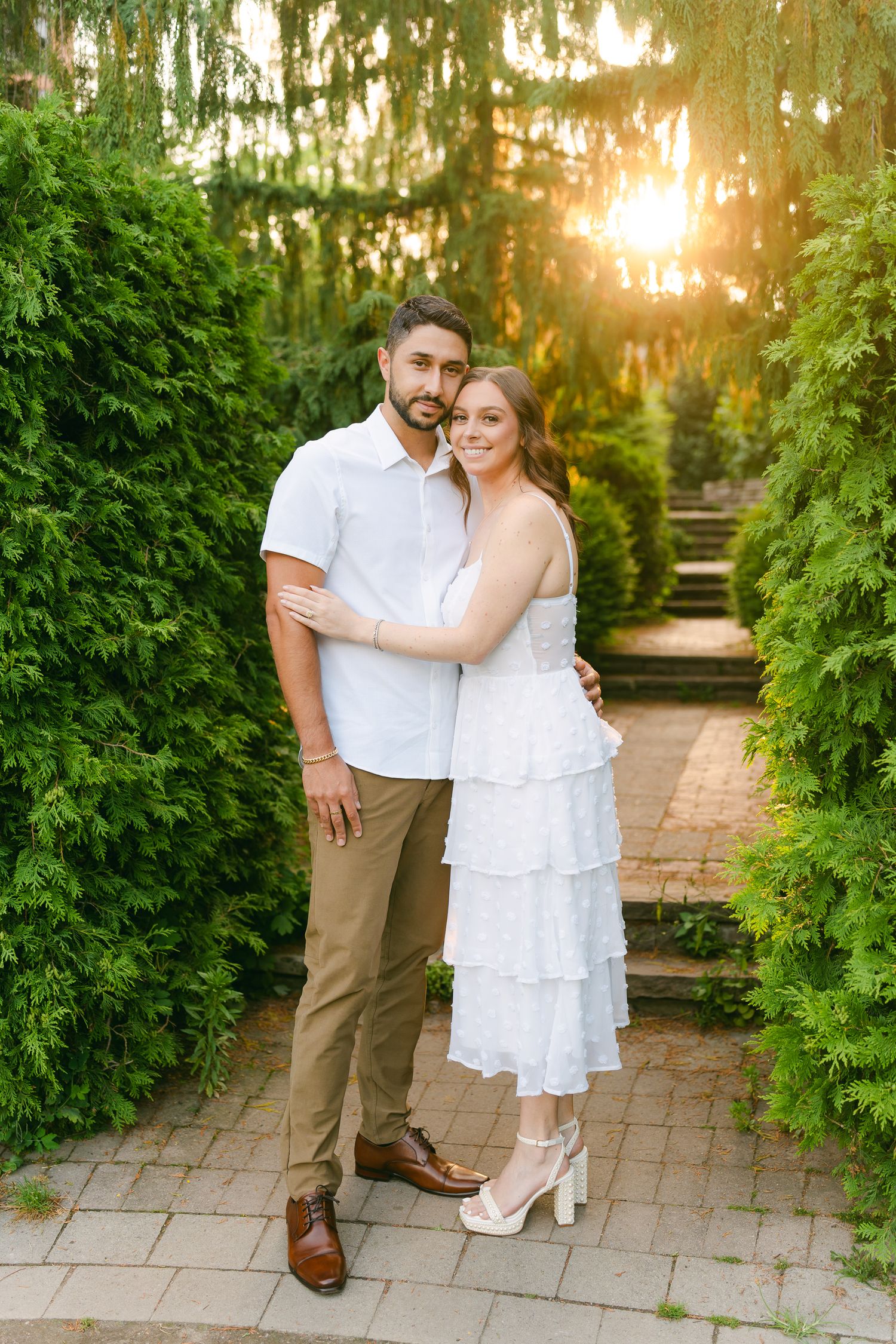 Couple in summer attire poses together on brick steps surrounded by green hedges.