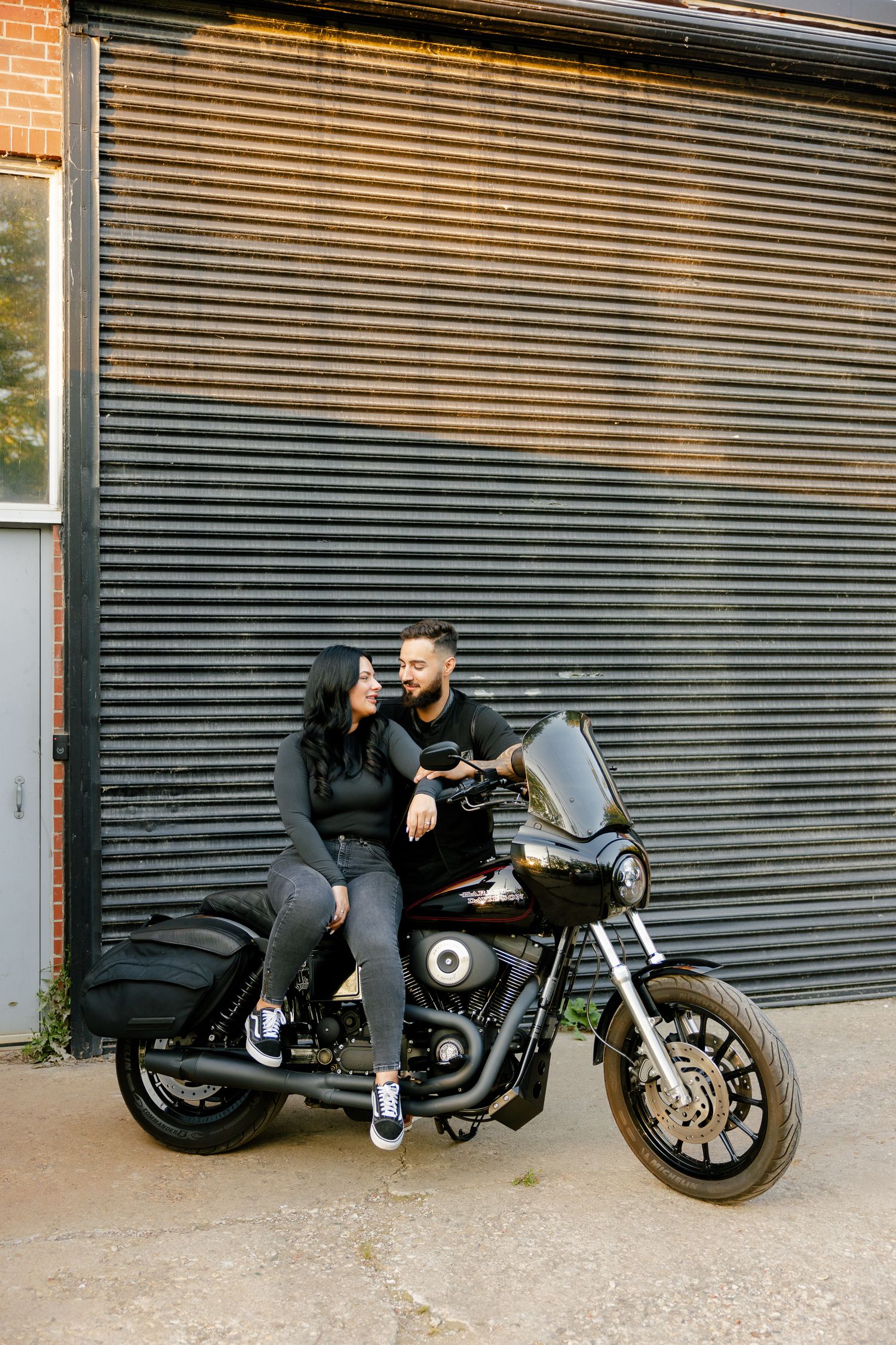 A couple embraces while seated on a parked motorcycle against an industrial wall.