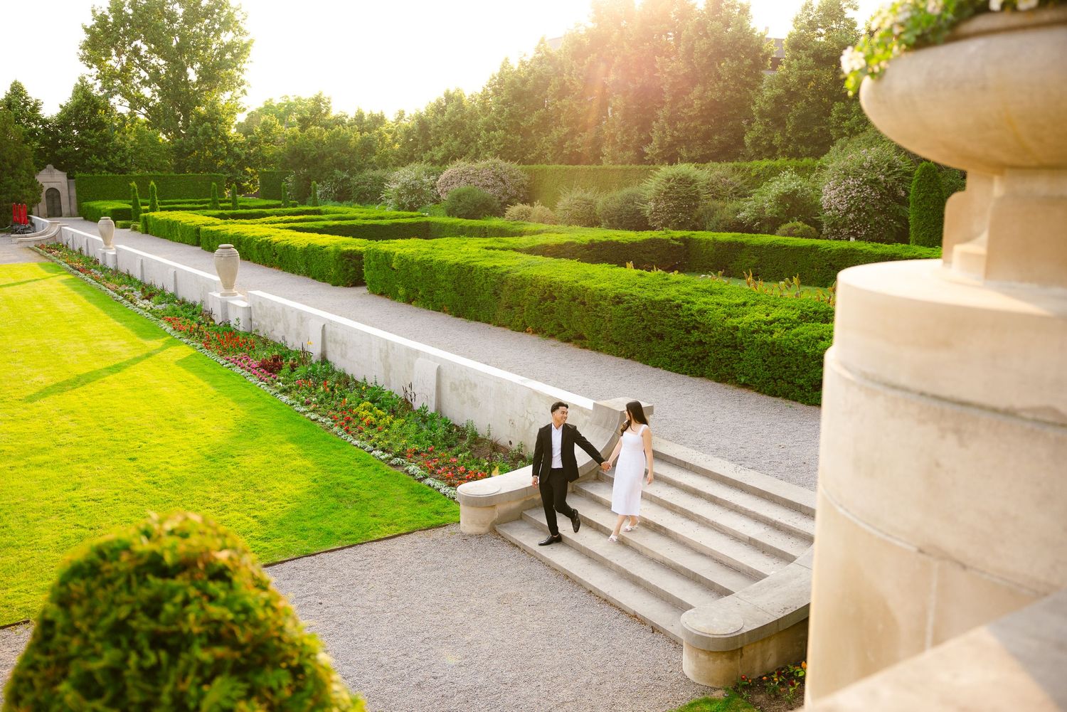 A couple walking down stone garden steps at sunset with manicured hedges and a decorative urn visible.