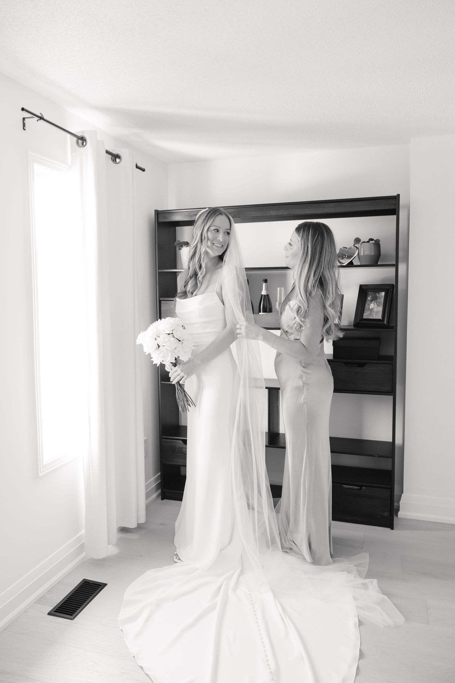 Two people in formal attire stand in a bright room with built-in shelves and a white wedding dress train.