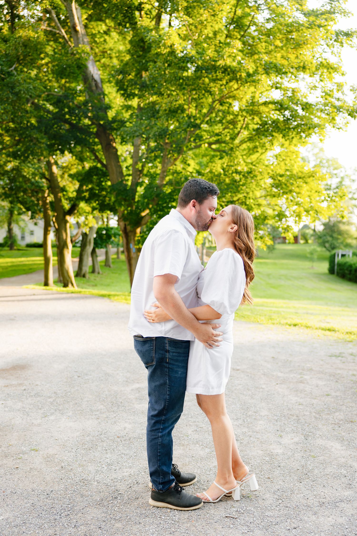 A romantic couple shares a tender moment on a sunlit path surrounded by lush green trees.