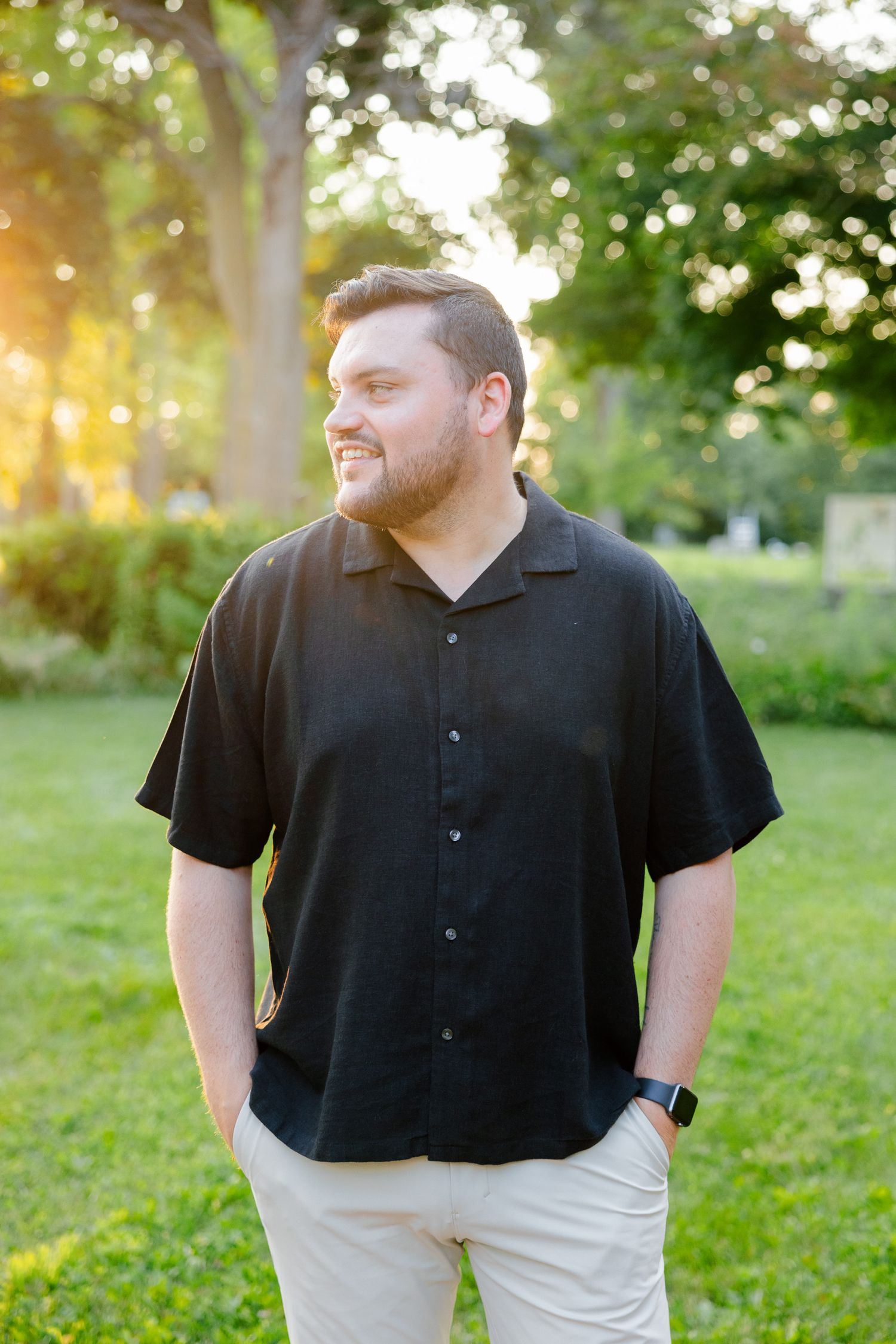 A person in a black button-down shirt smiles while standing in a garden during golden hour.