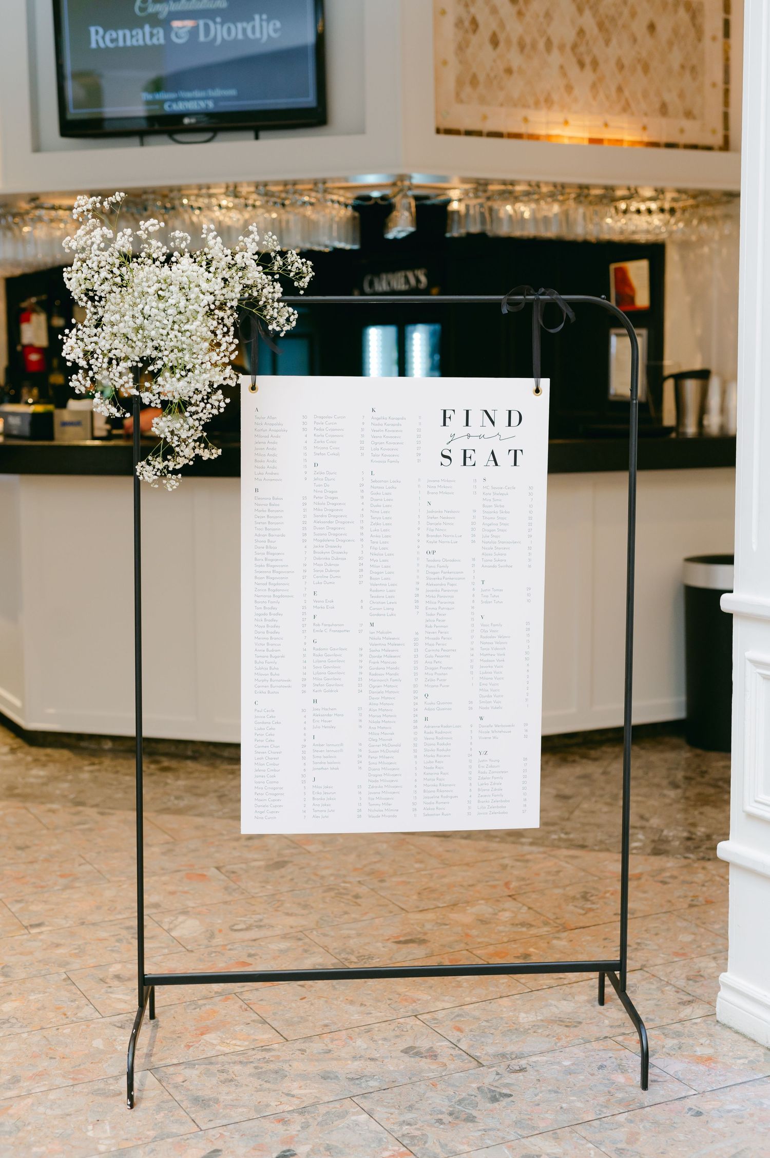 Wedding seating chart display board adorned with delicate baby's breath flowers on a black metal stand.