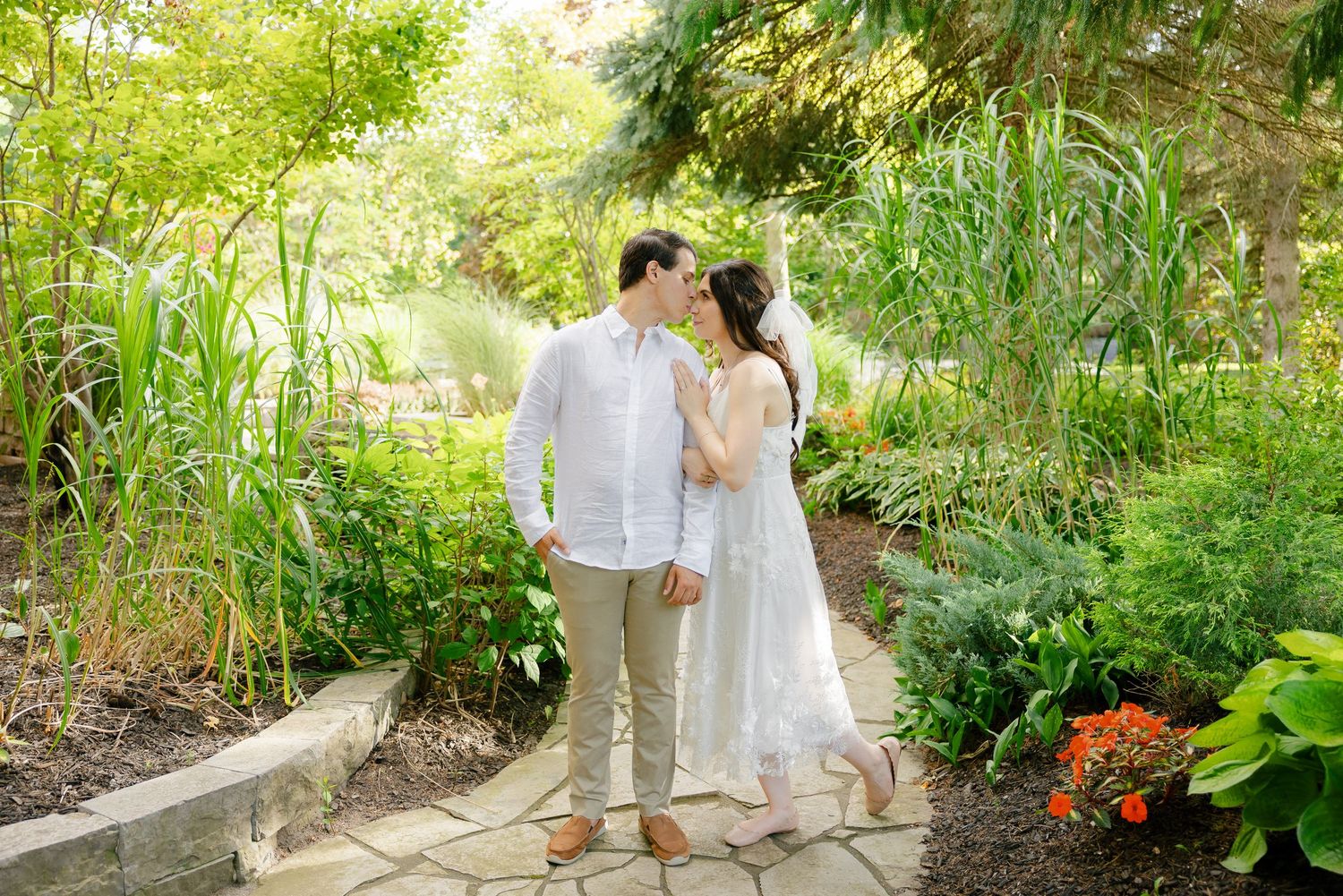 A couple embraces on a stone pathway surrounded by lush summer gardens.