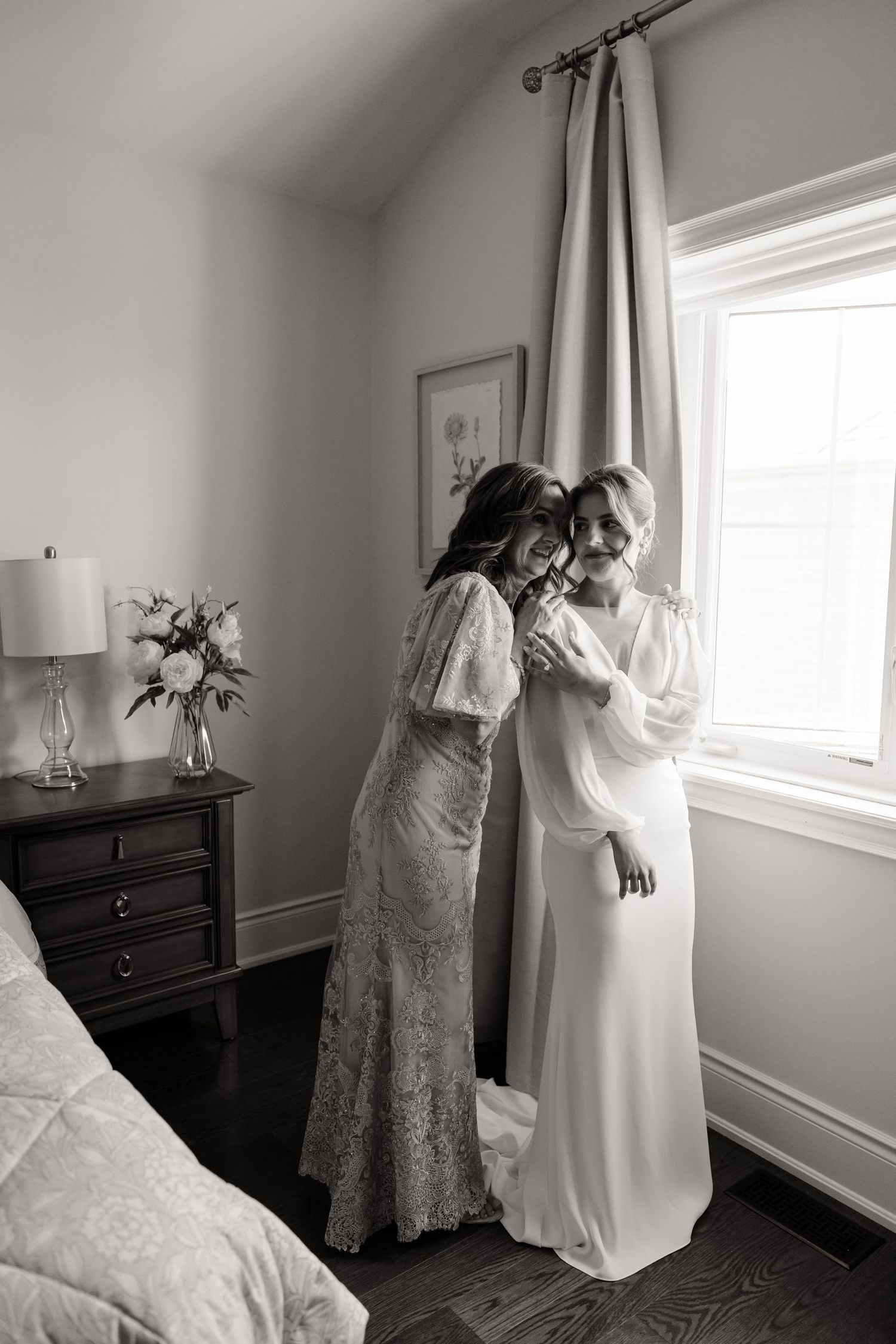 A black and white photo shows two people in elegant dresses sharing a tender moment by a window.