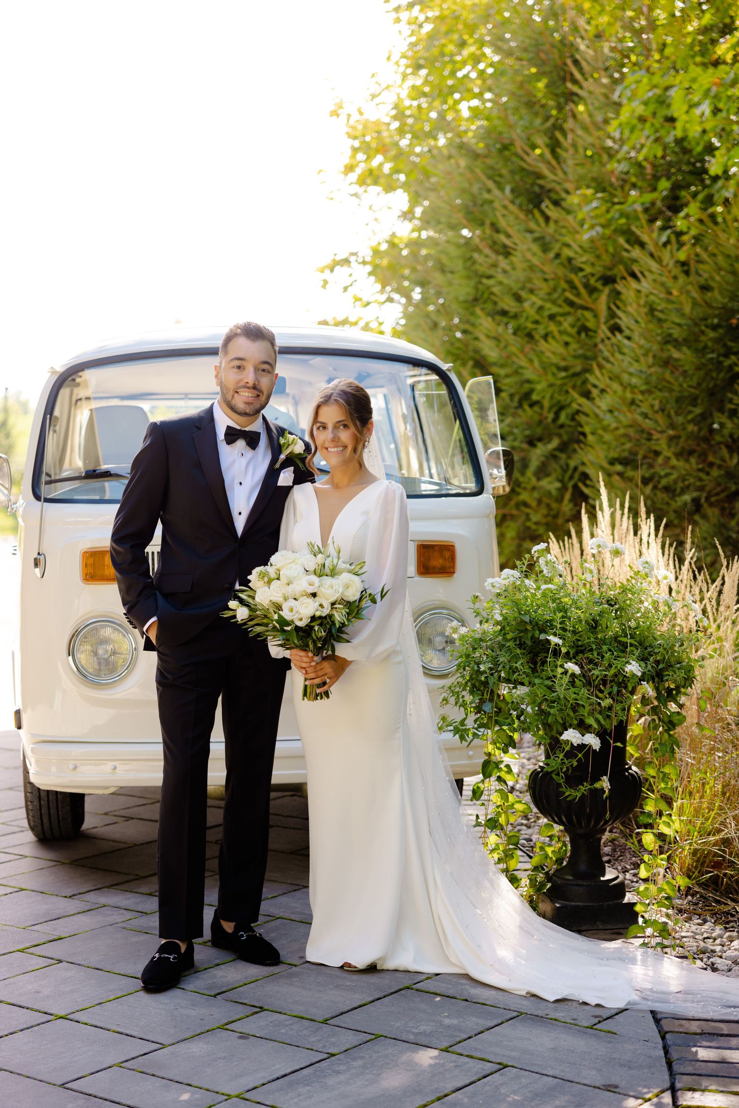Newlyweds pose in formal attire next to a vintage white VW van surrounded by lush greenery.