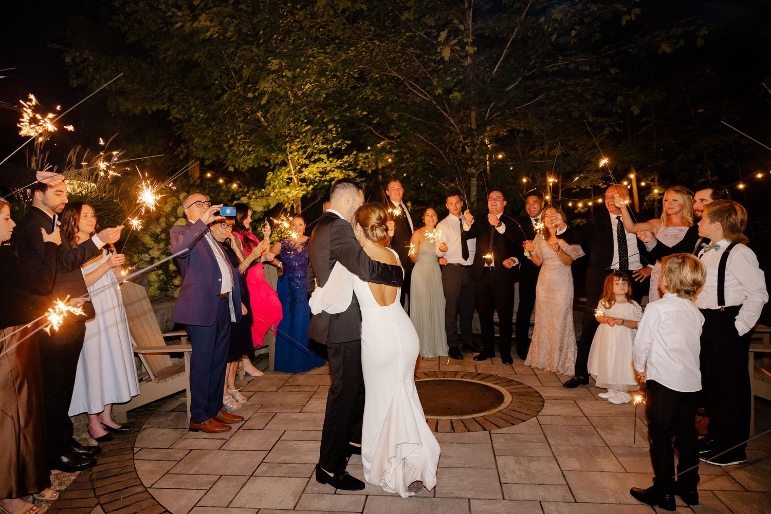 Wedding guests holding sparklers form a circle around the dancing couple on a stone patio.