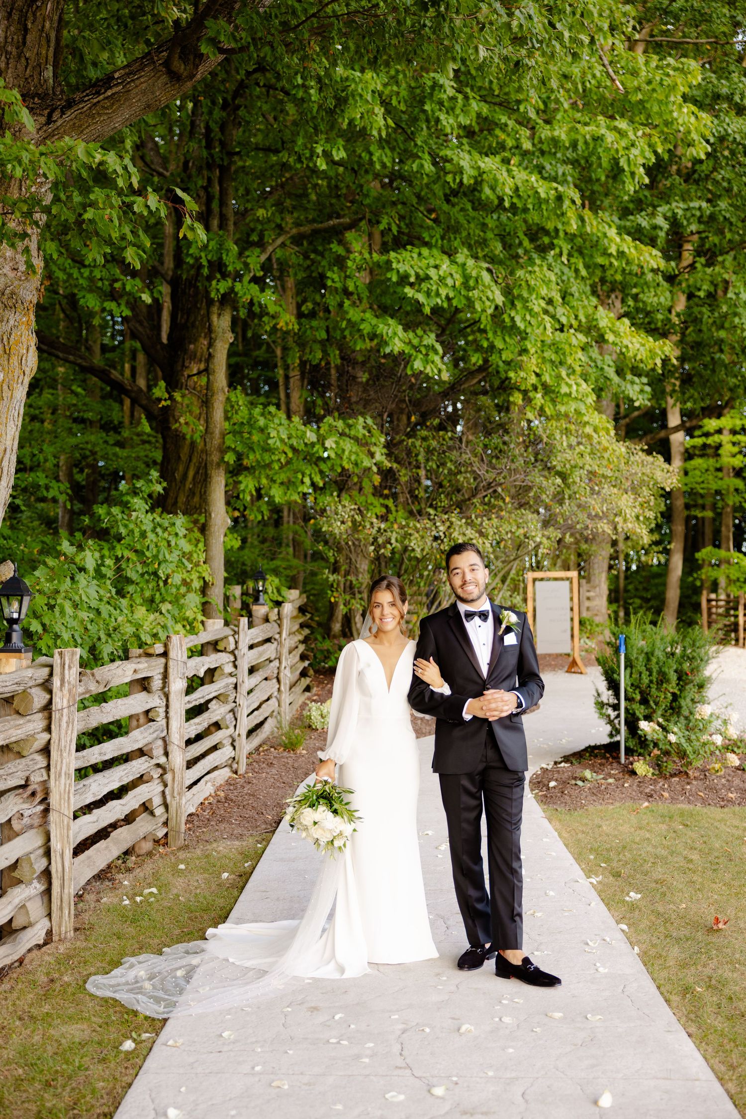A wedding couple poses on a pathway surrounded by lush green trees and wooden fencing.