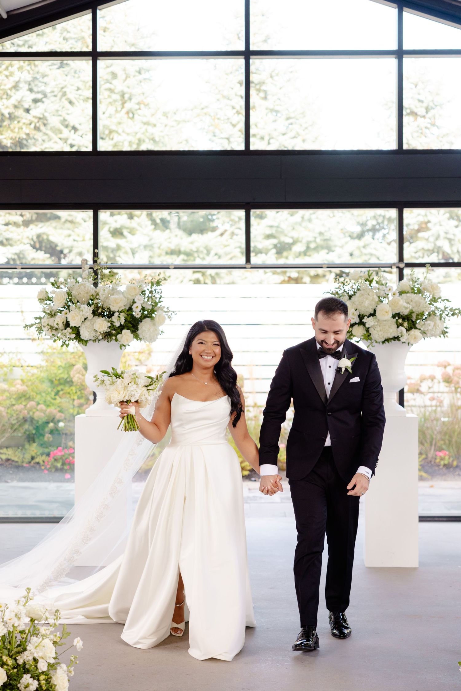 A newlywed couple walks together in a modern venue decorated with elegant white floral arrangements.