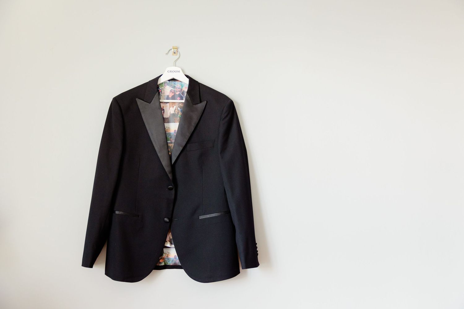 A black tuxedo jacket hangs on a white hanger against a plain wall.