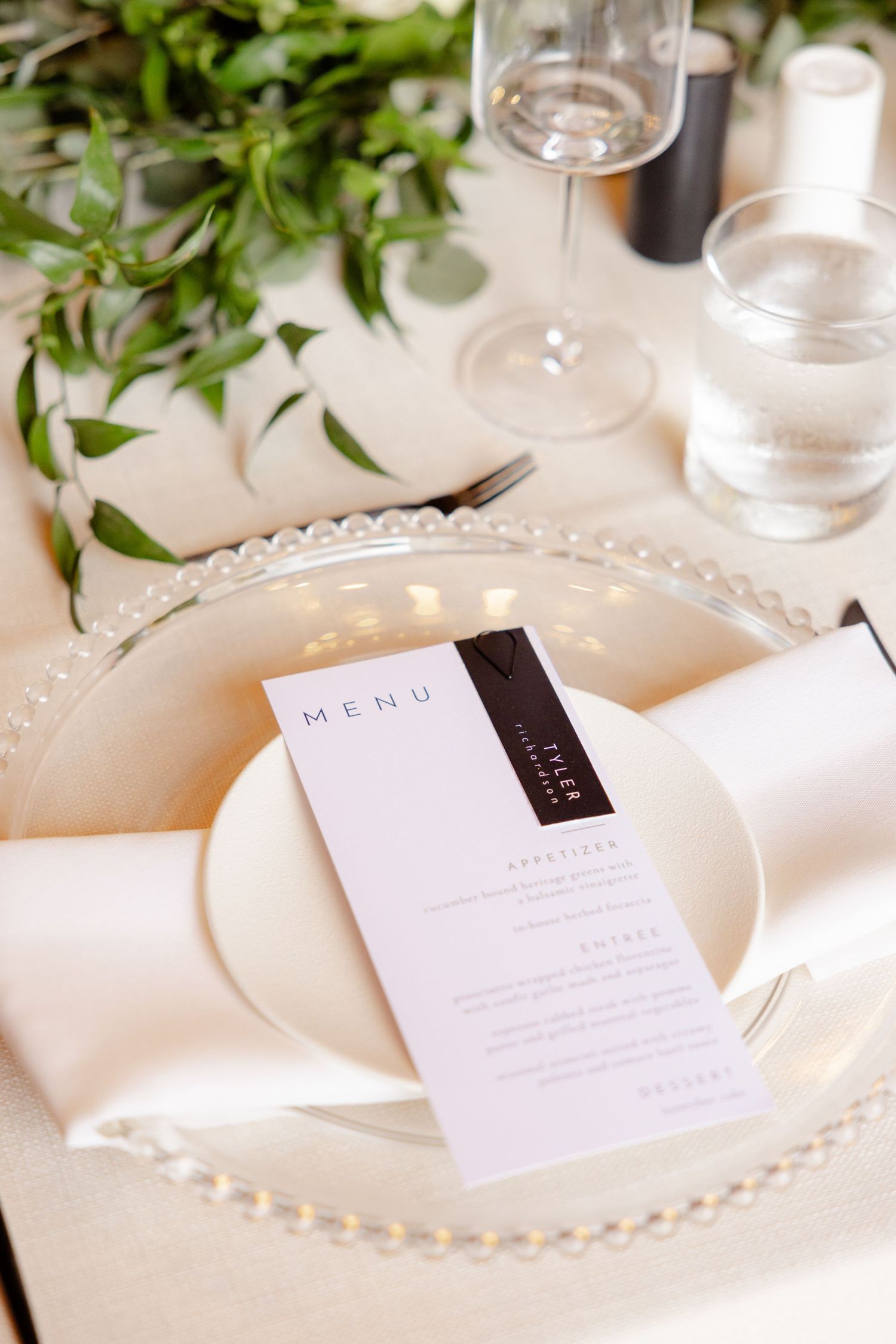 Wedding place setting with elegant menu card on beaded charger plate surrounded by greenery.