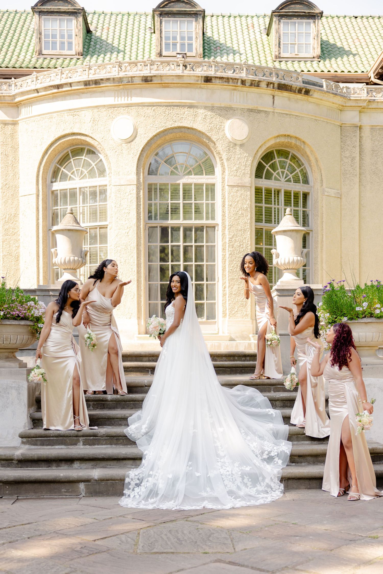 A wedding party in champagne dresses poses playfully on stone steps of a historic venue.