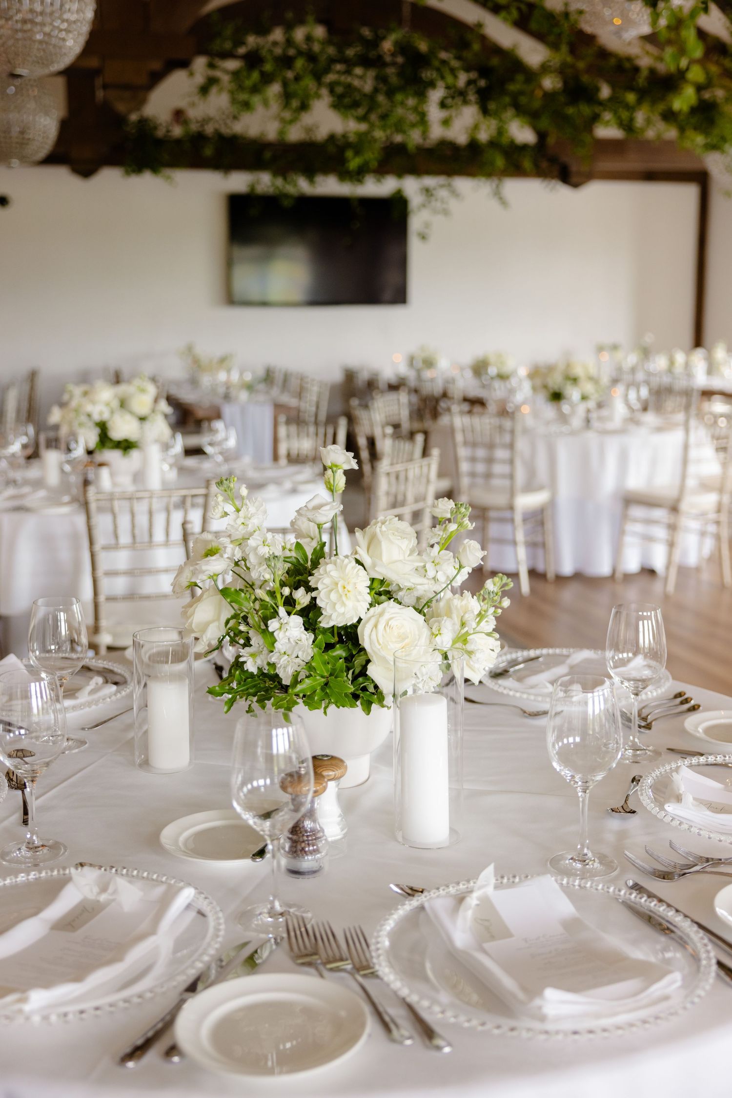 Round wedding reception tables with white linens, floral centerpieces, and crystal glassware under wood beams.