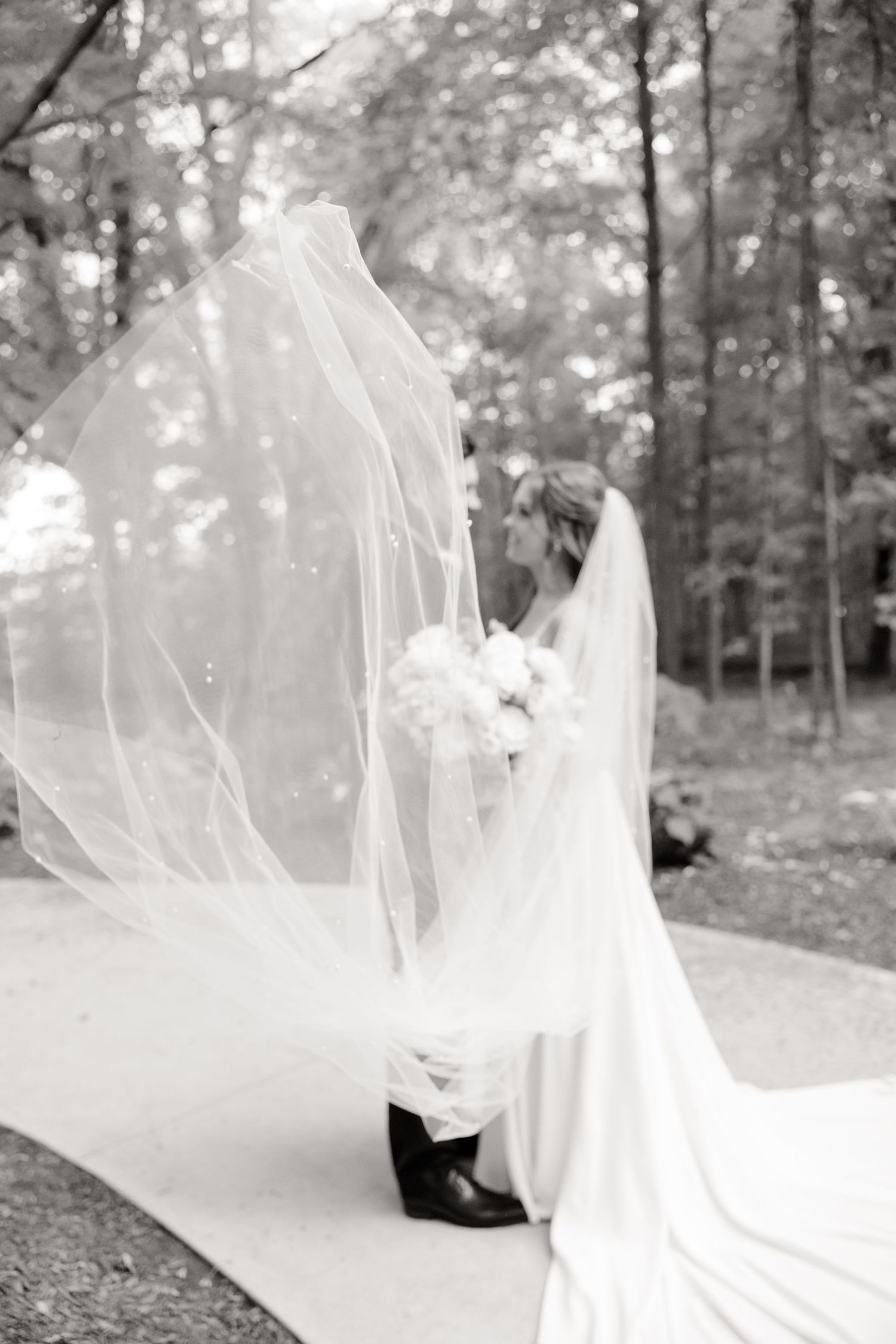 Black and white wedding photo showing flowing veil movement against forest backdrop.