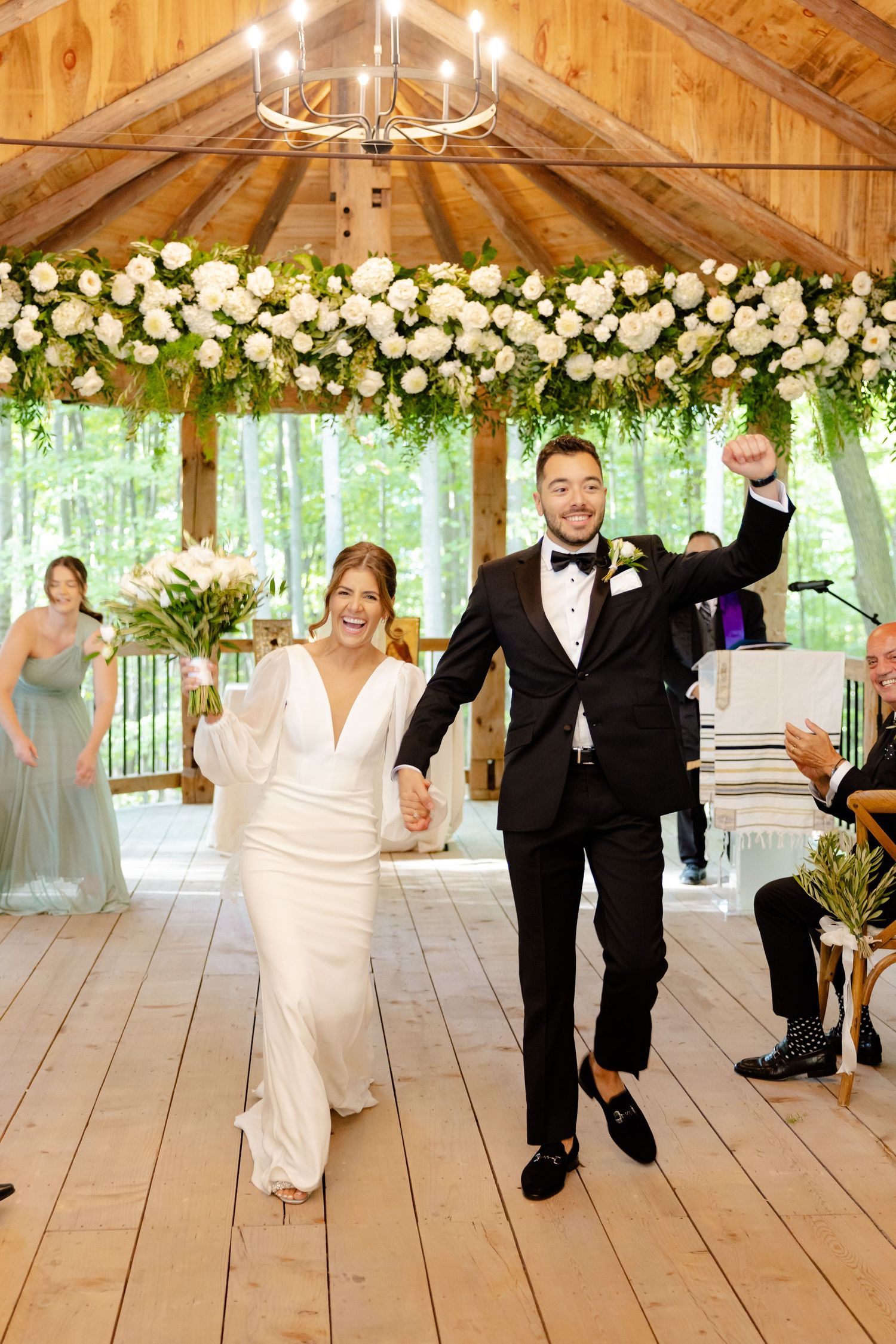 Newlyweds celebrate their exit while walking down the aisle in a wooden pavilion with floral decorations.