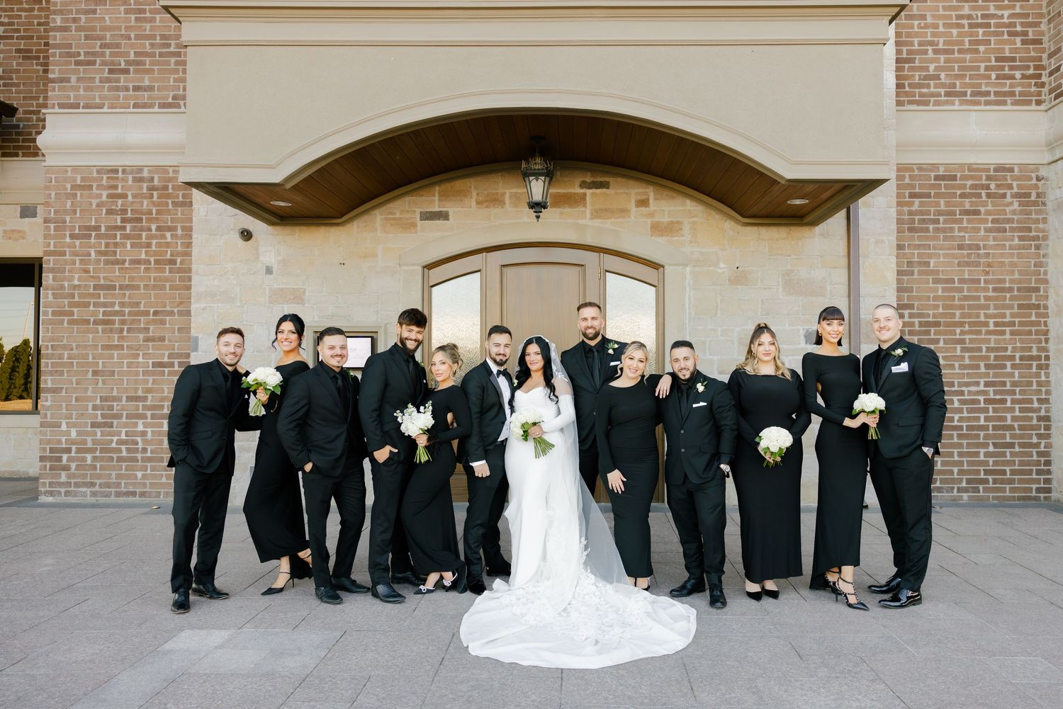 Wedding party in classic black attire poses with bride in white gown against architectural backdrop.