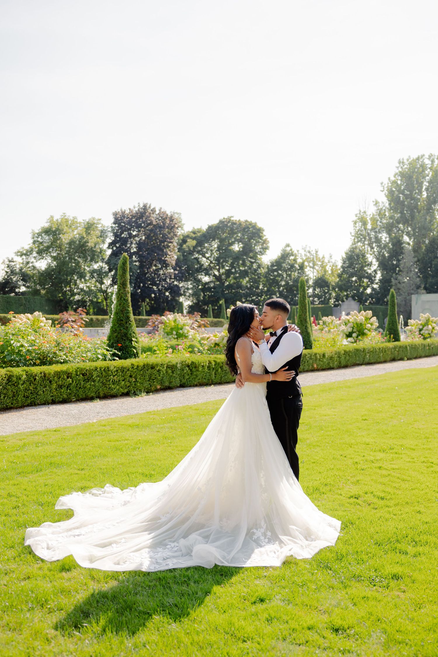 Sunlit garden wedding portrait featuring a couple in classic attire among topiary and flowers.