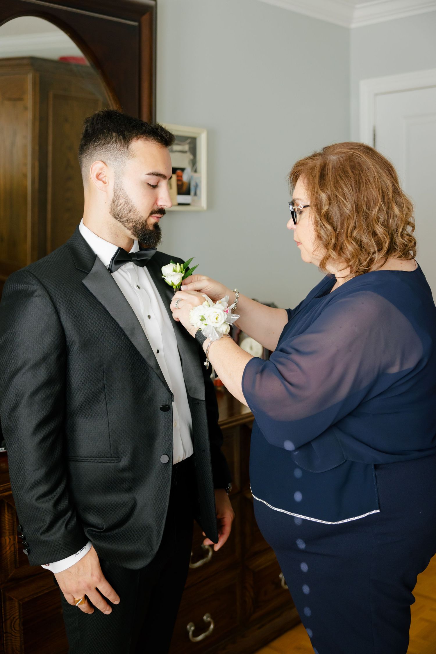A woman in navy blue helps pin a white boutonniere onto a groom's black tuxedo lapel.