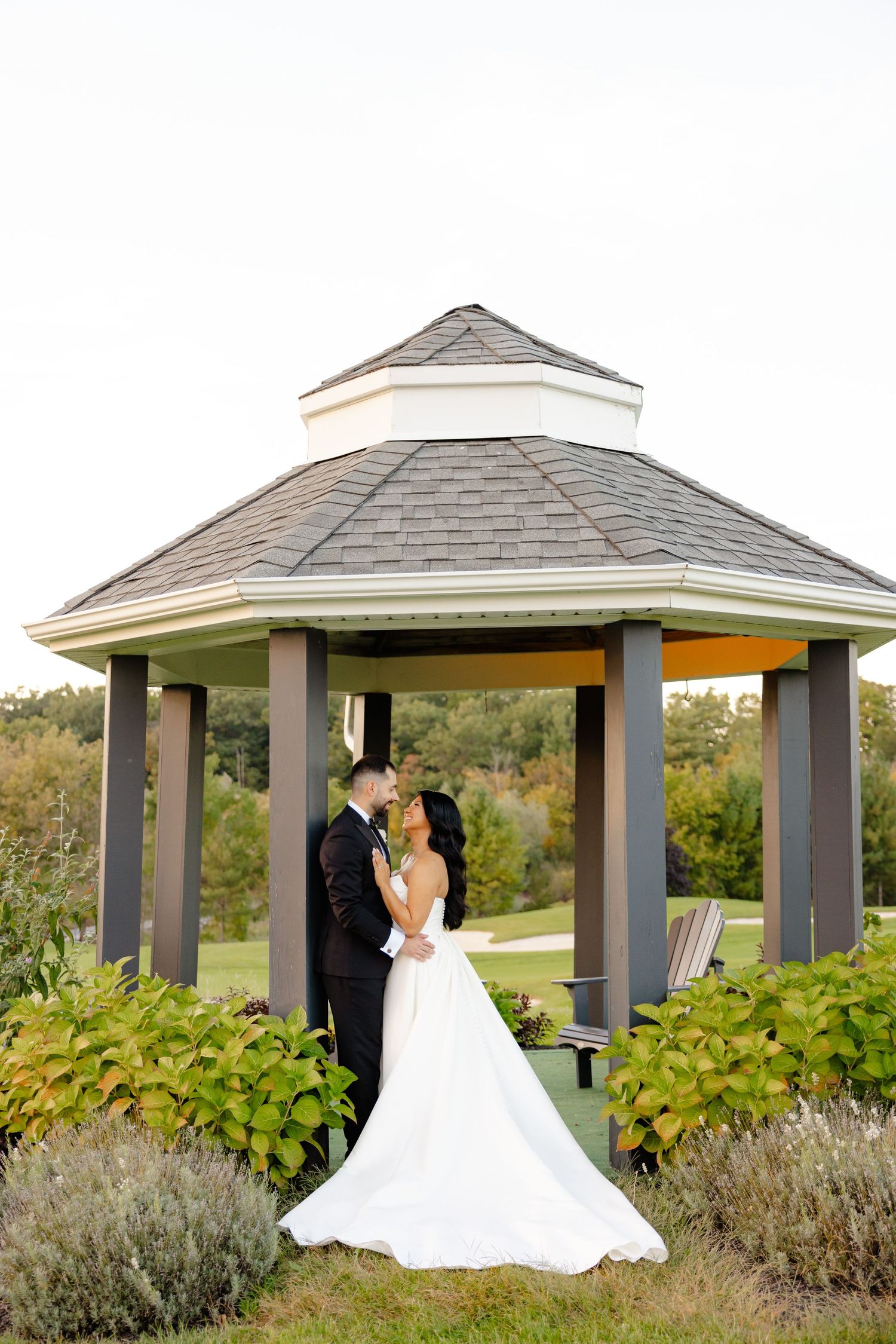 A wedding couple shares an intimate moment under a gray hexagonal gazebo surrounded by lush greenery.