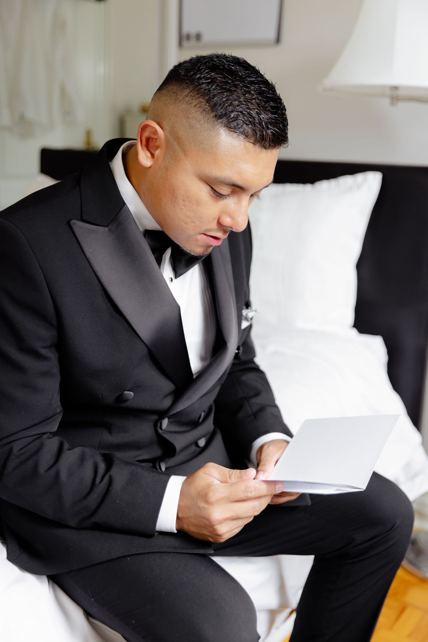 Man in black tuxedo sits reading wedding vows from paper during wedding preparation.