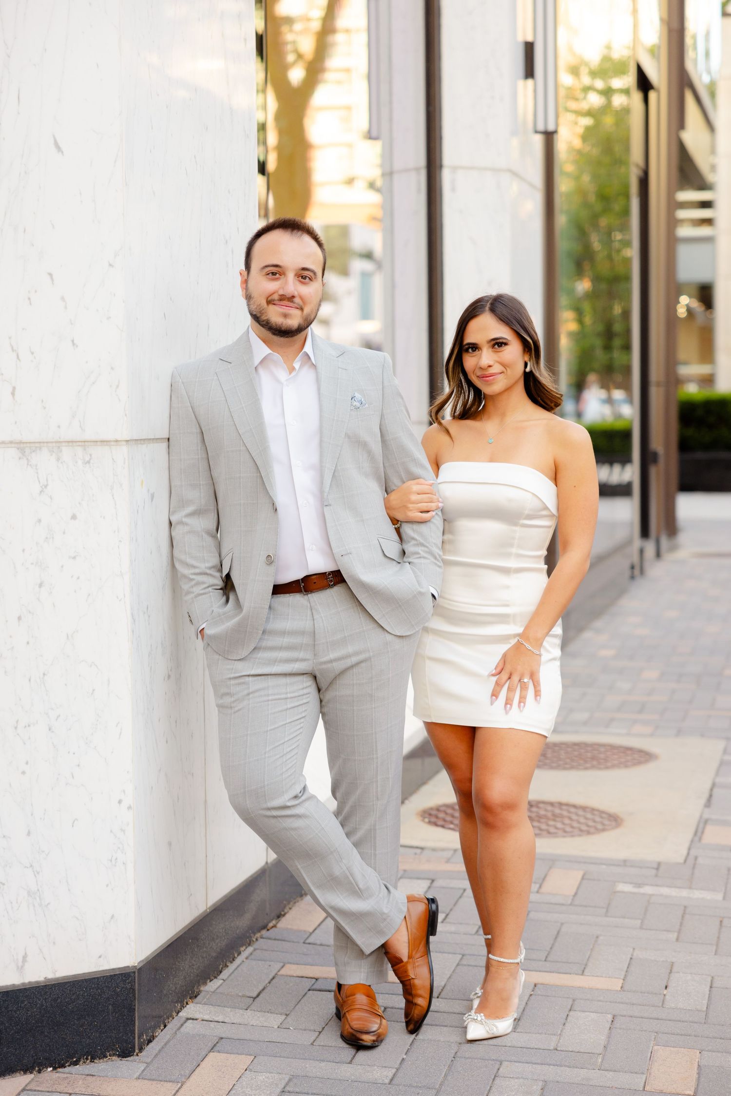 Fashionable couple in gray suit and white dress pose together against building exterior.