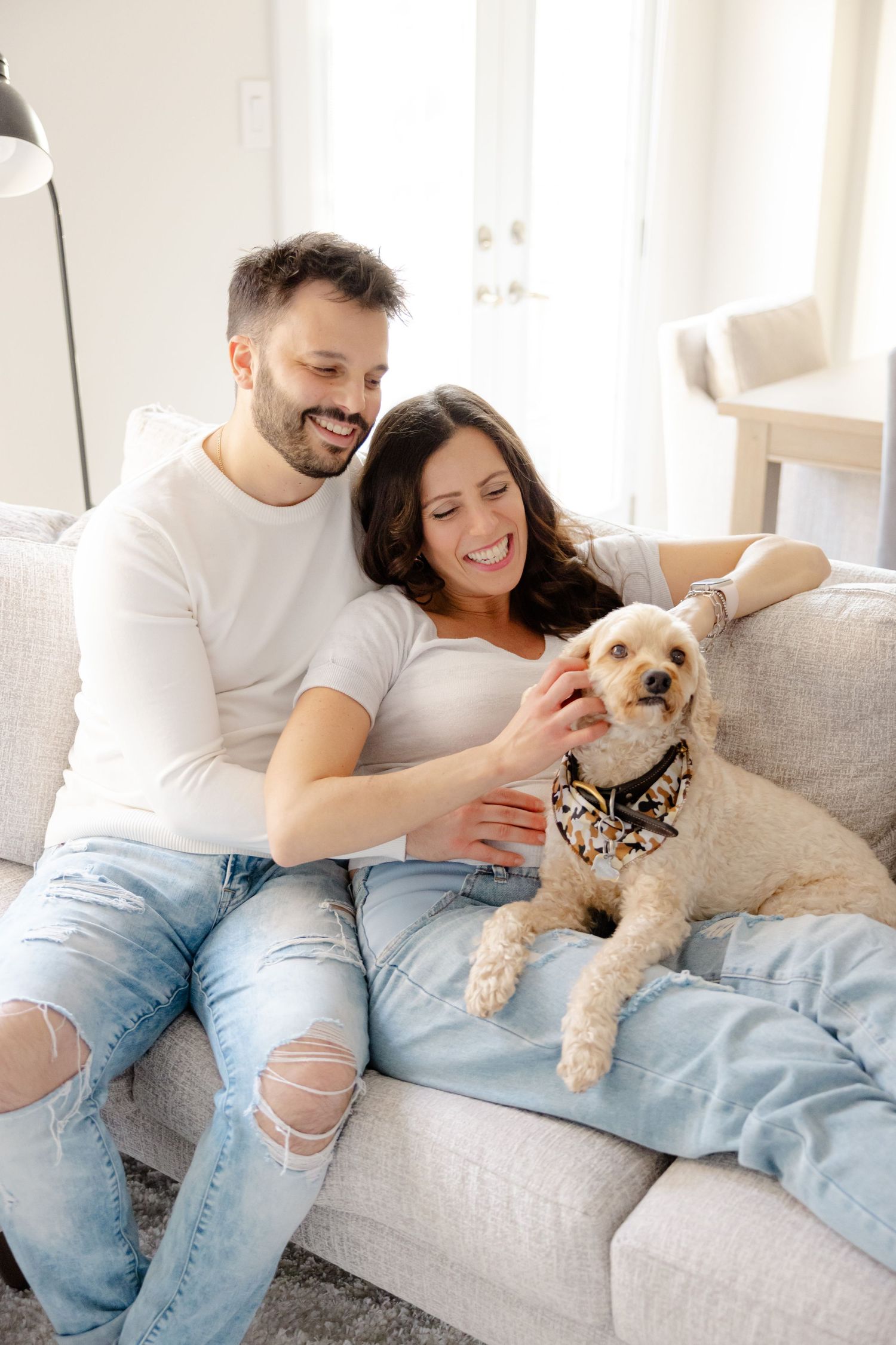 A couple sits on a couch with a small white dog wearing a bandana, all smiling in a bright room.
