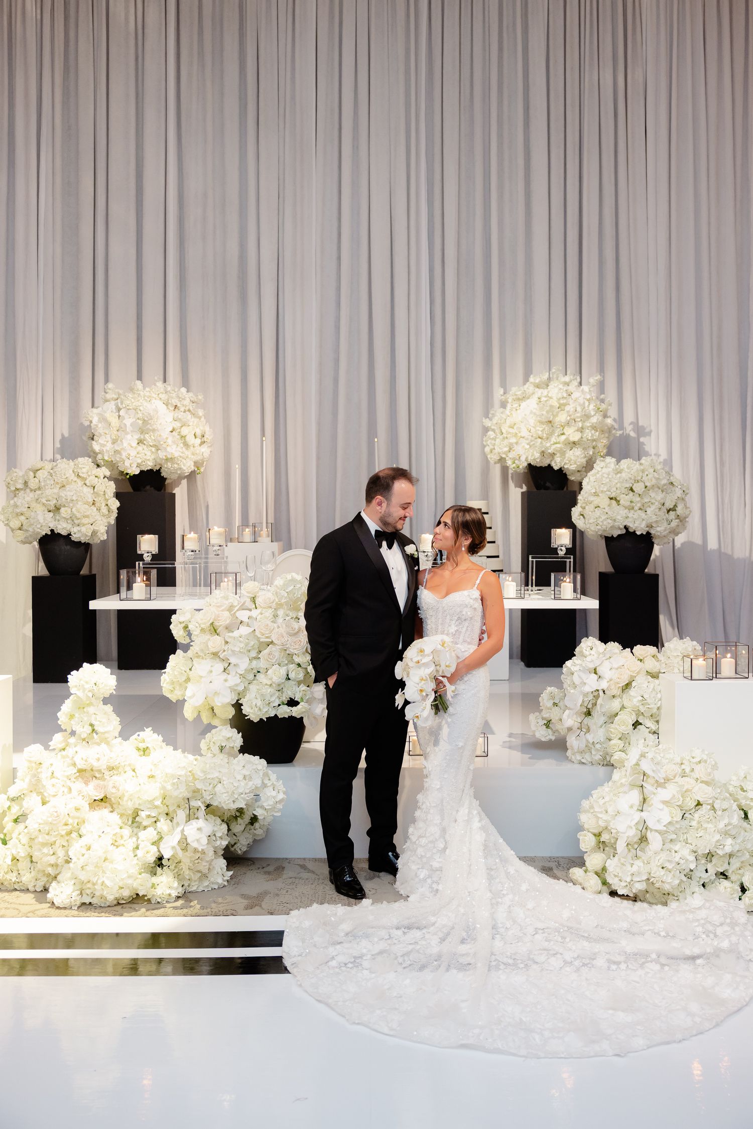 Elegant wedding scene with couple surrounded by luxurious white floral arrangements and floor-to-ceiling drapes.