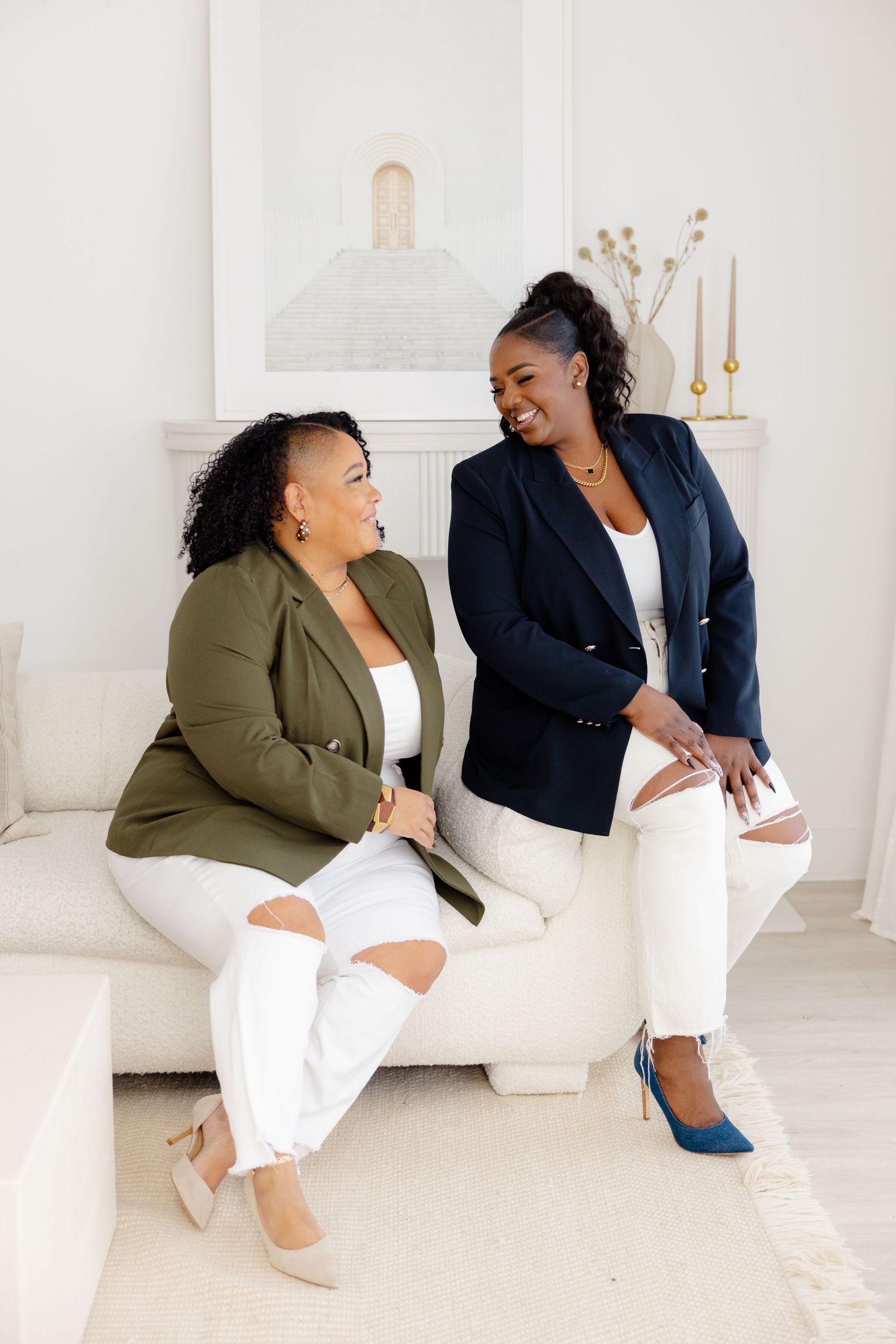 Two women in business attire share a laugh while sitting on a white couch in a bright, modern space.