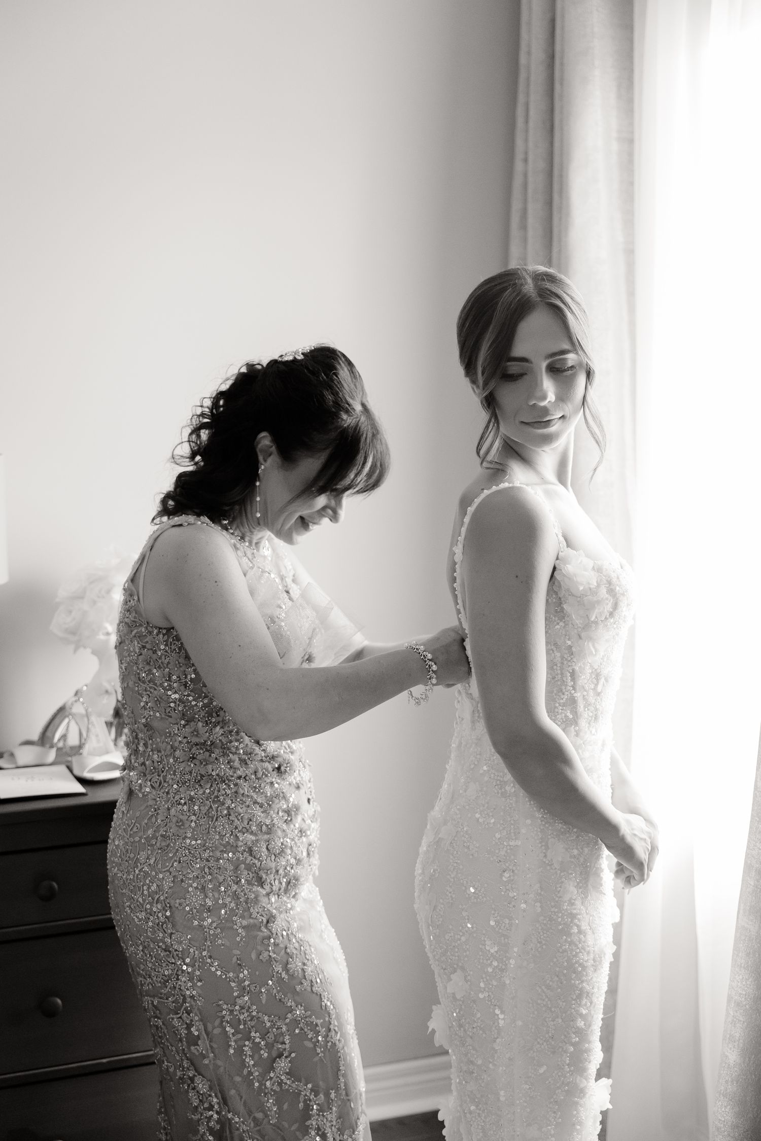 Final preparations of a bride getting into her sparkling white wedding dress in a sunlit dressing room.
