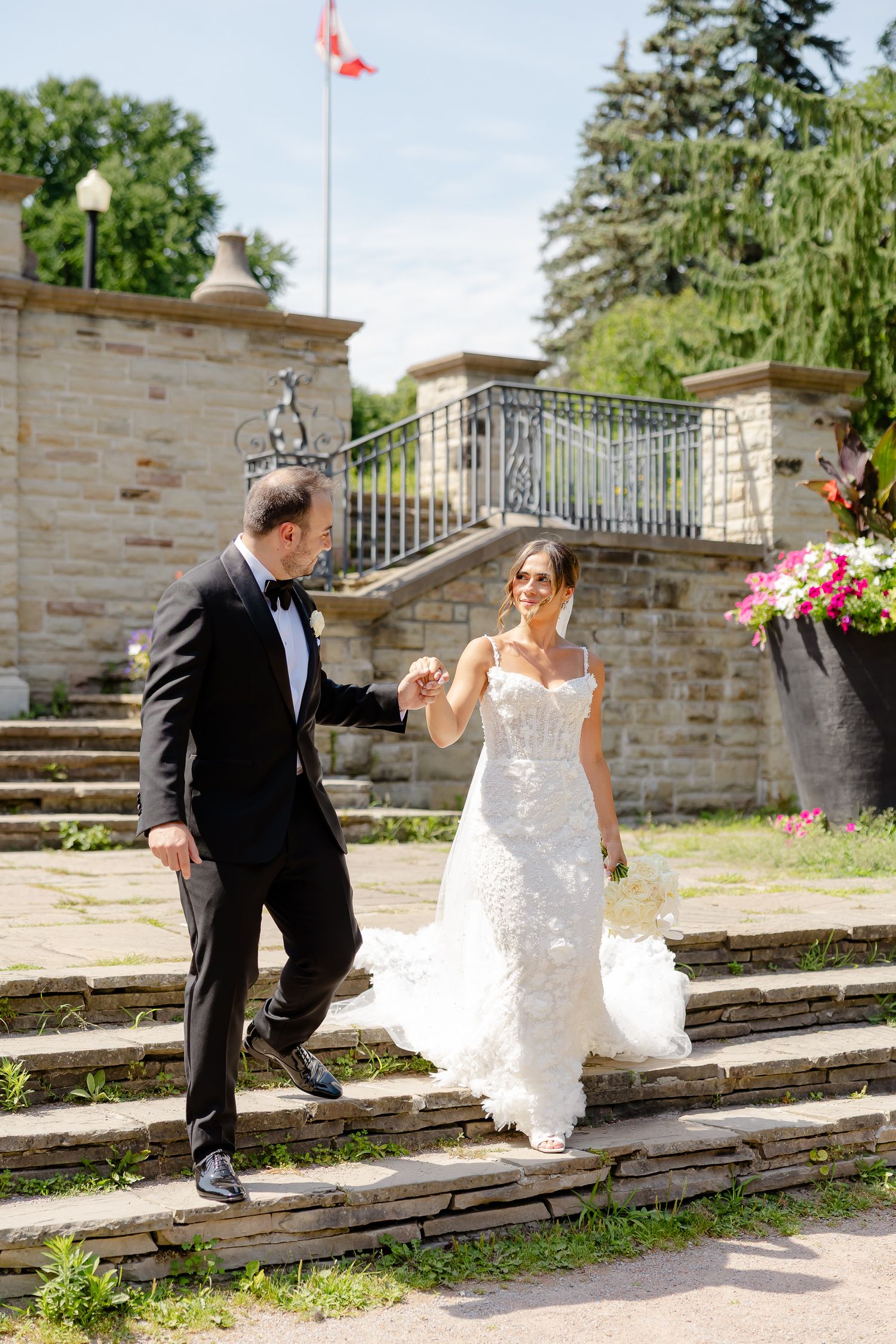 A couple in wedding attire playfully spins and dances down stone steps in a garden setting.