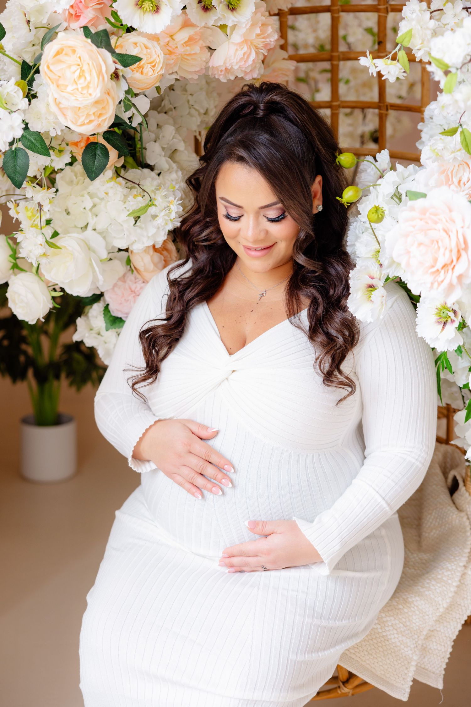 Expecting mother in white knit dress poses near floral arch with peach roses and white hydrangeas during maternity photoshoot.