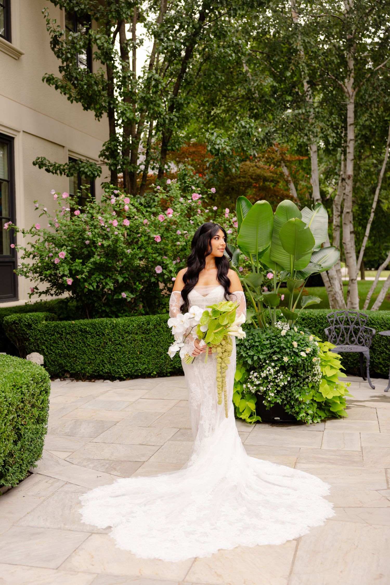 Bride in white lace gown with long train poses in garden courtyard surrounded by manicured hedges.