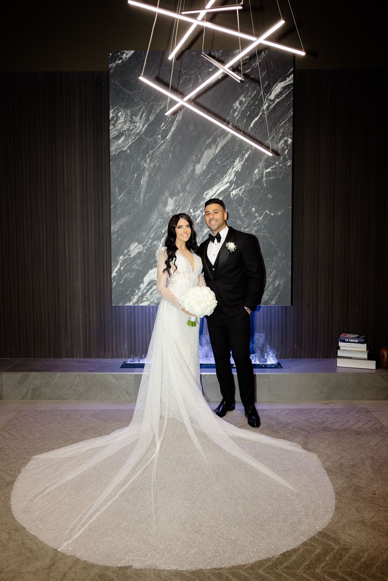 Bride and groom stand under modern lighting fixture against marble backdrop at wedding venue.