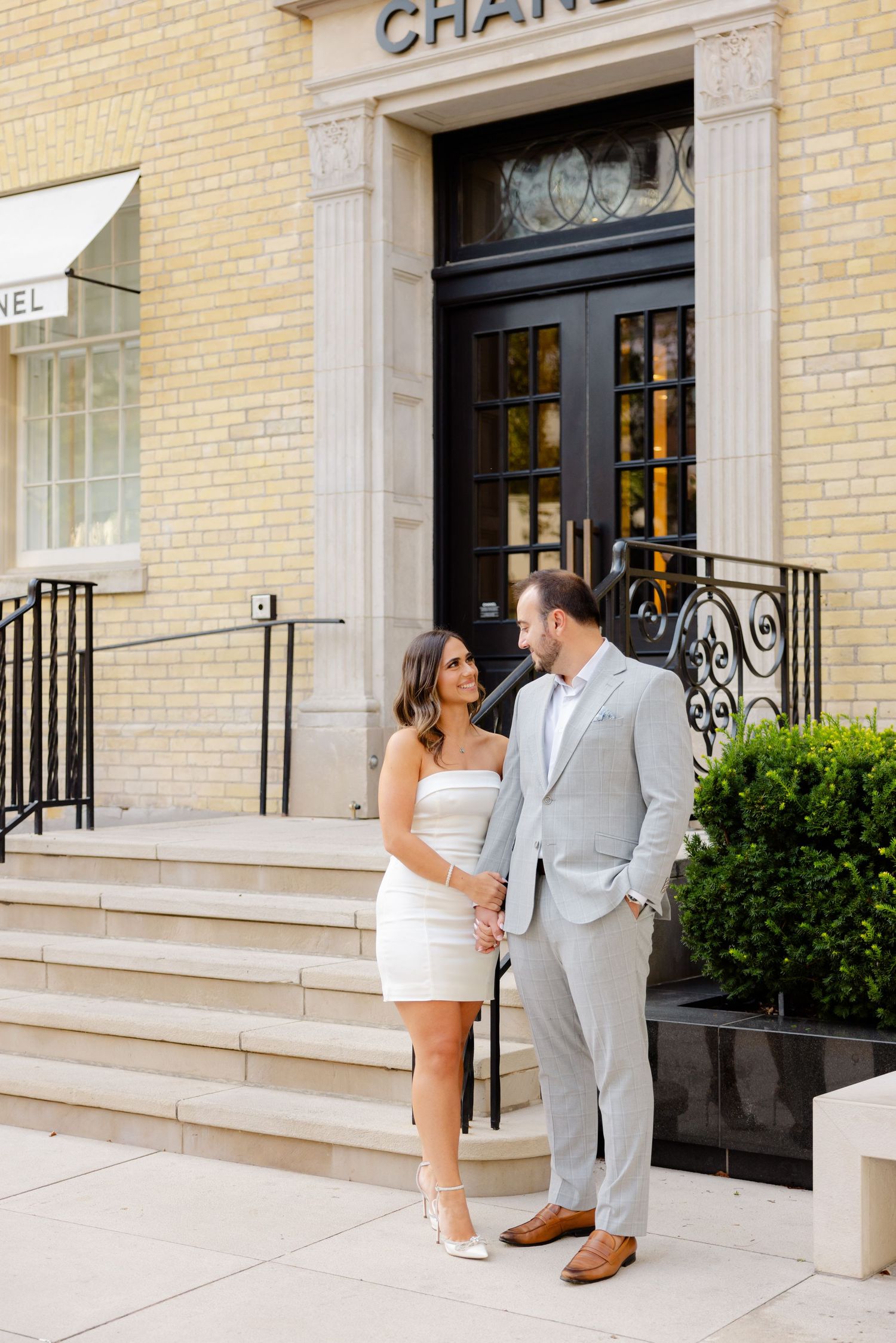 Elegant couple stands together on marble steps of Chanel boutique with classic storefront design.
