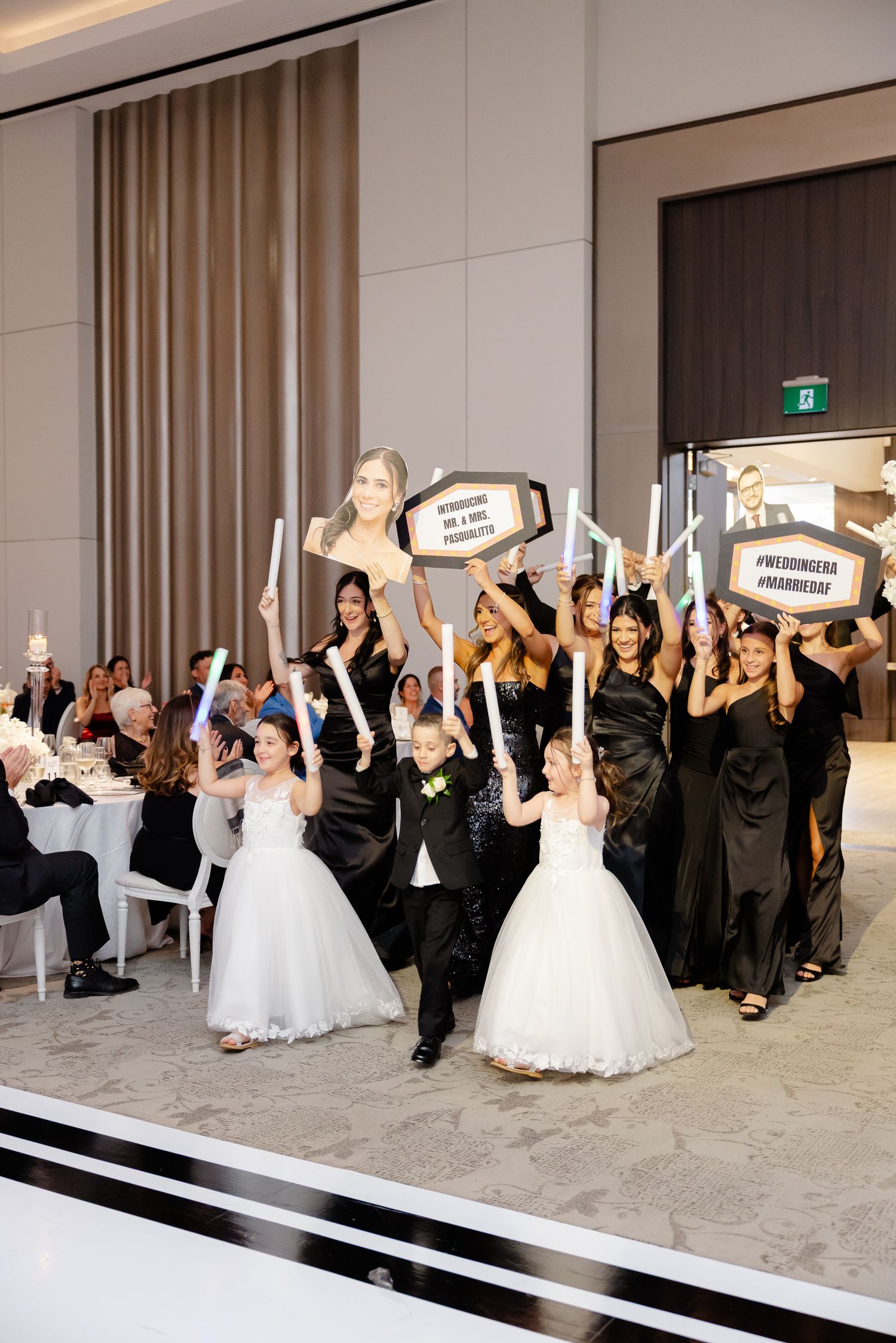 Wedding party celebrates on stage with fun props and signs while wearing black dresses and white gowns at elegant reception.