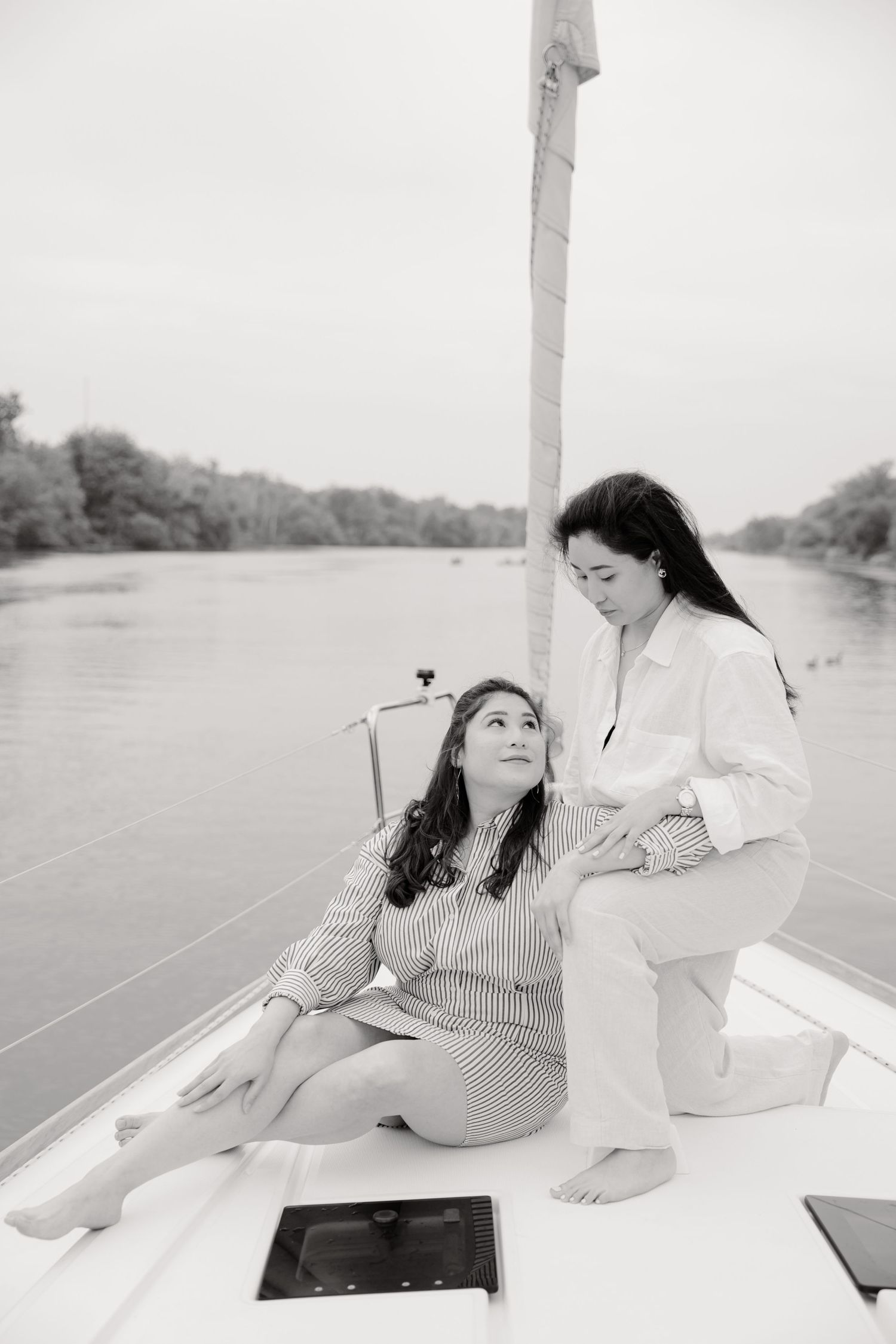 Two women relax together on a boat deck near a calm river, captured in black and white photography.