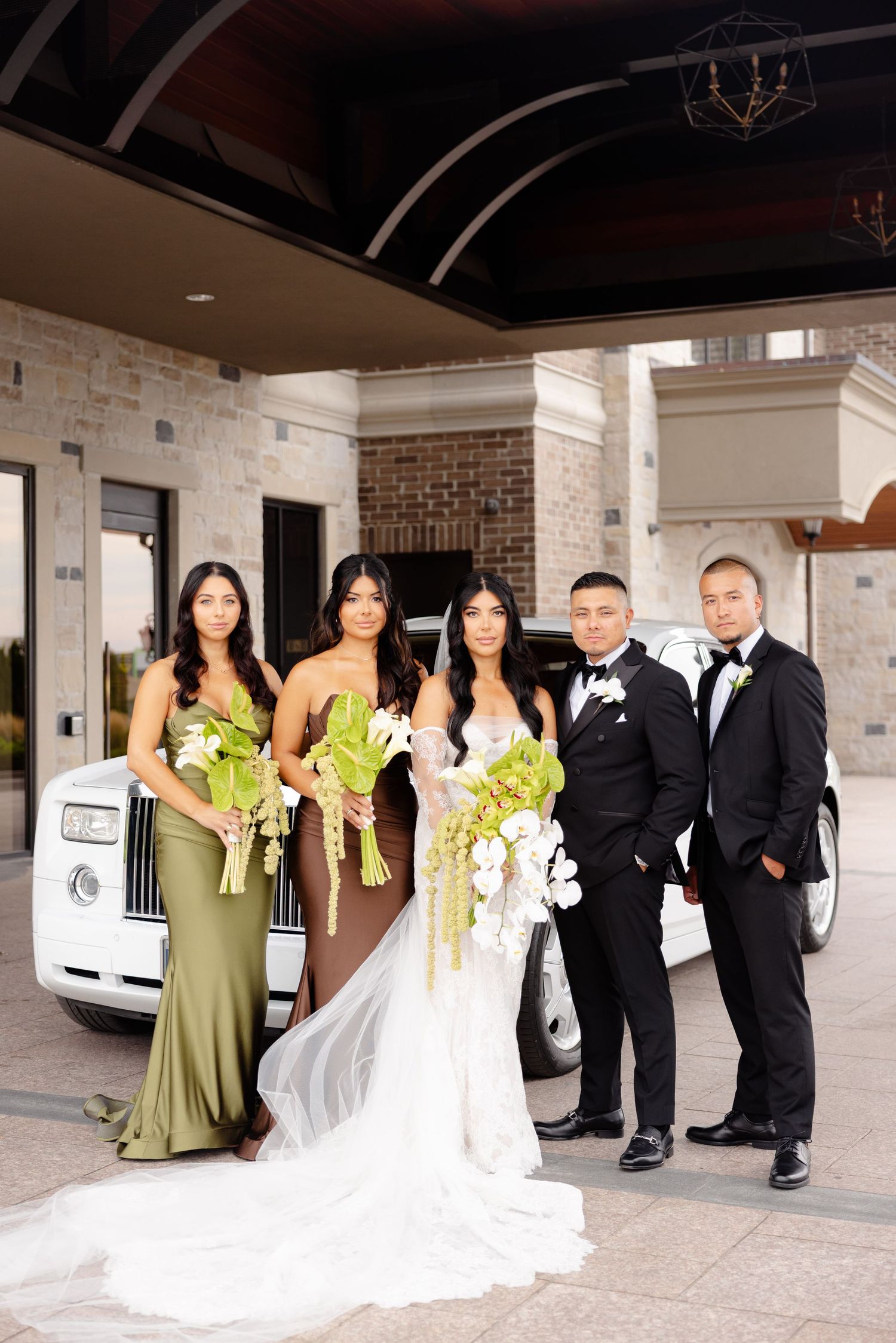 Wedding party poses with white cars and green bouquets by stone building entrance.