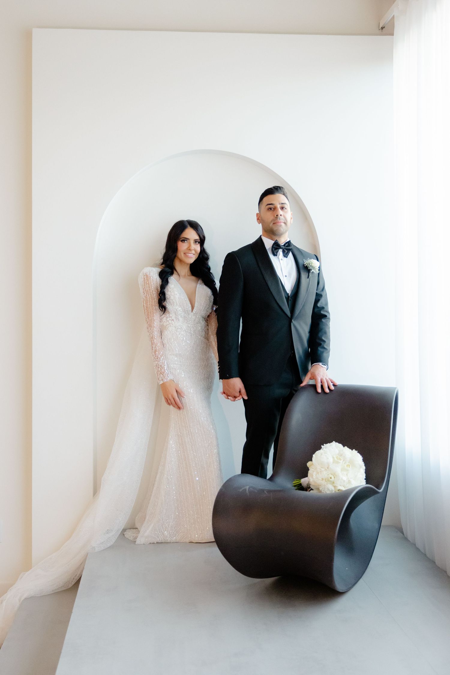 Modern wedding portrait with couple standing in white archway next to contemporary brown chair.