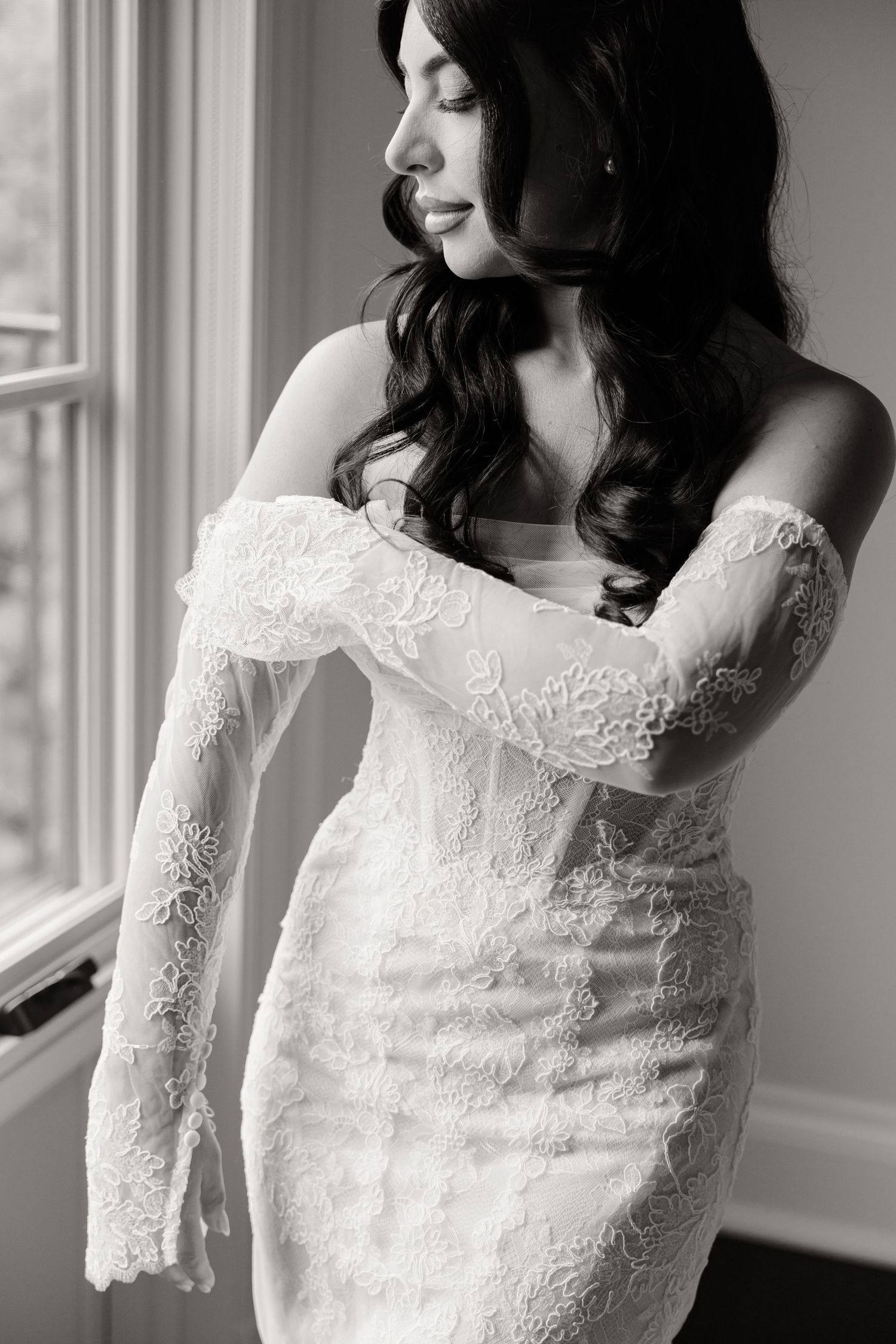 Black and white portrait of a bride wearing a fitted lace wedding gown with long sleeves near a window.