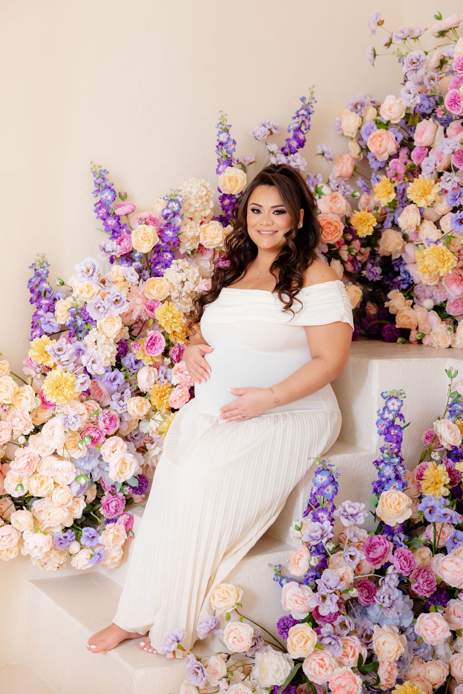 Beautiful maternity portrait with off-shoulder white gown surrounded by pink, purple and yellow floral arrangements.