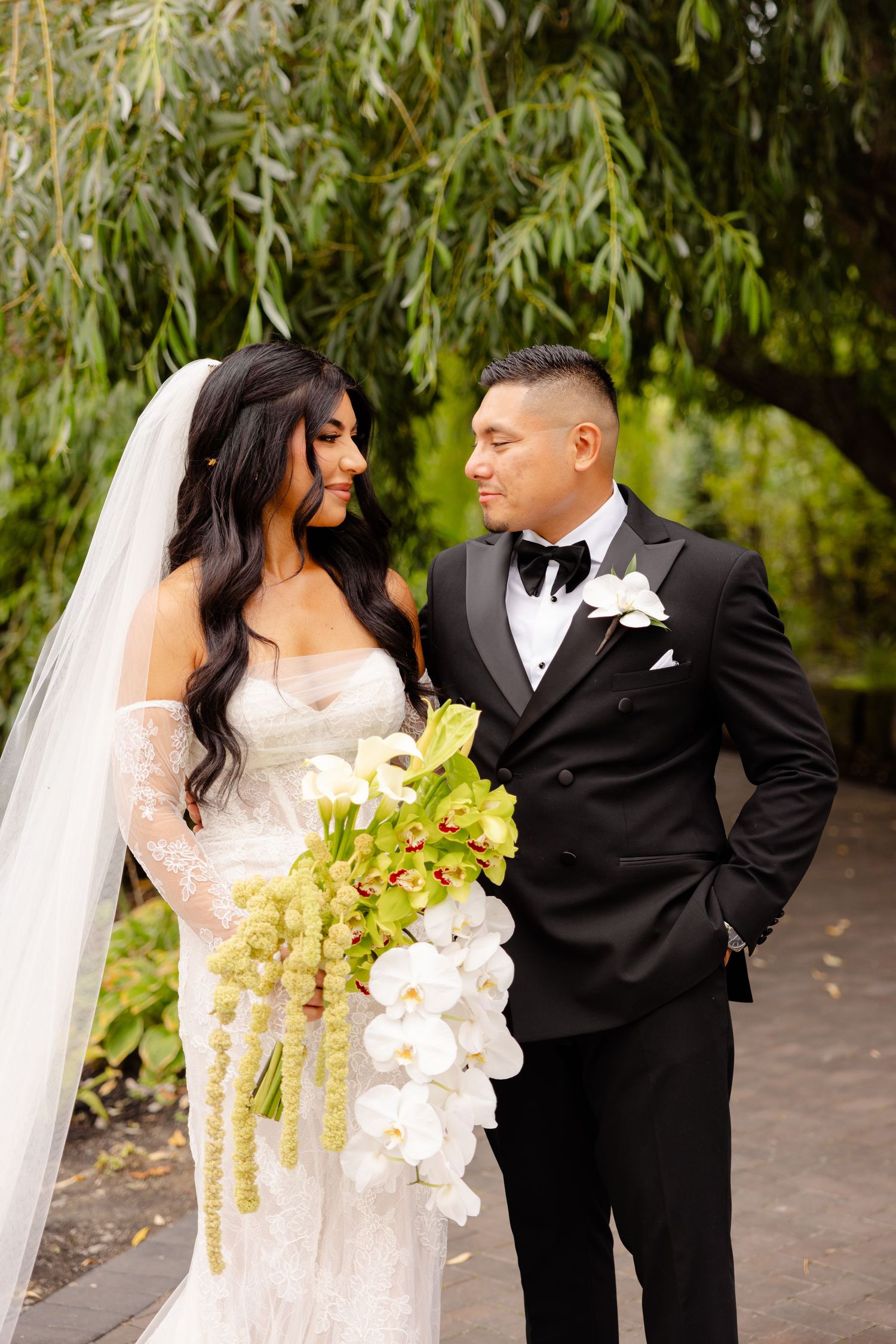 A bride in a white lace gown and groom in black tuxedo pose outdoors under willow trees with an orchid bouquet.
