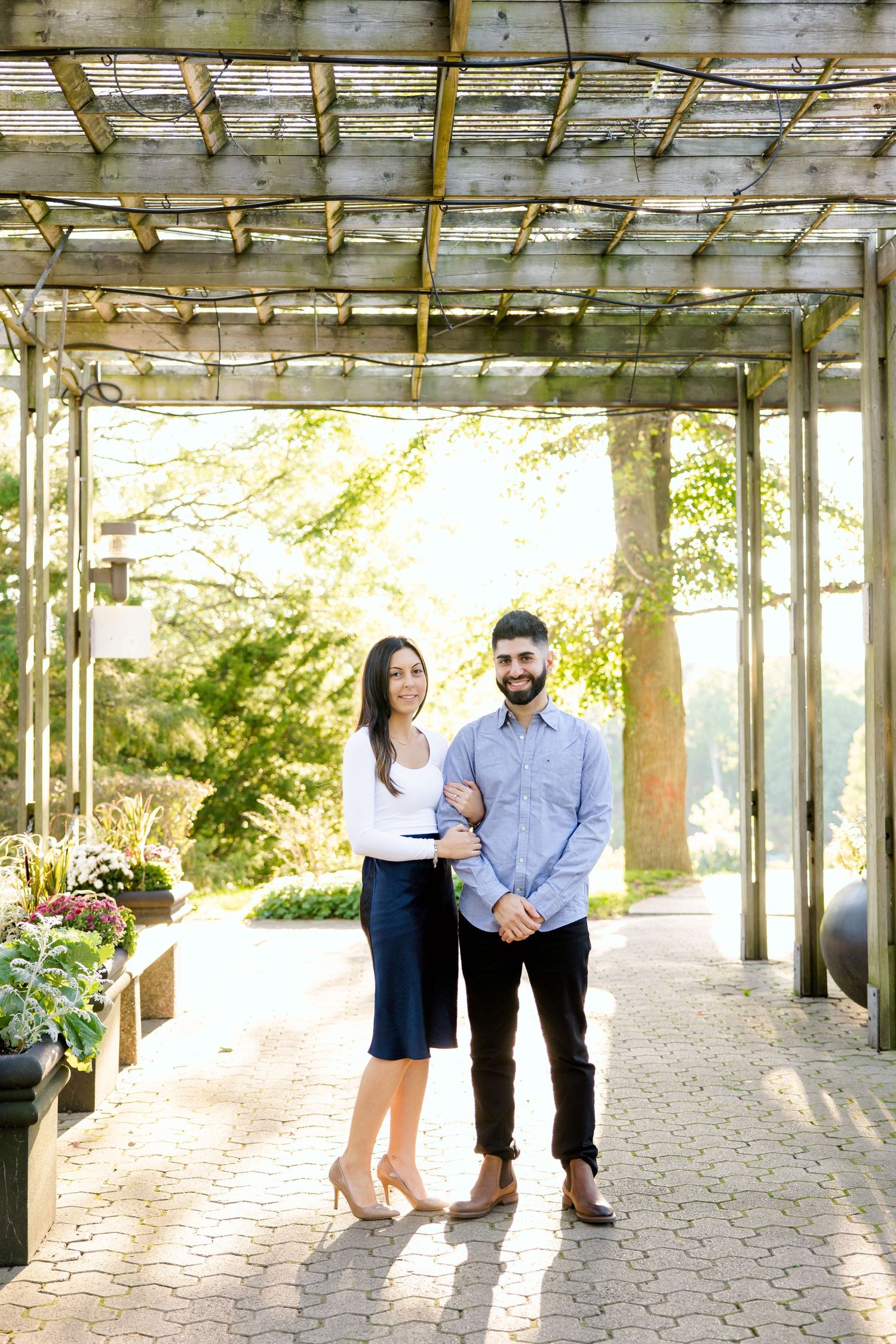 A couple poses beneath a garden pergola with sunlight streaming through on a brick pathway.